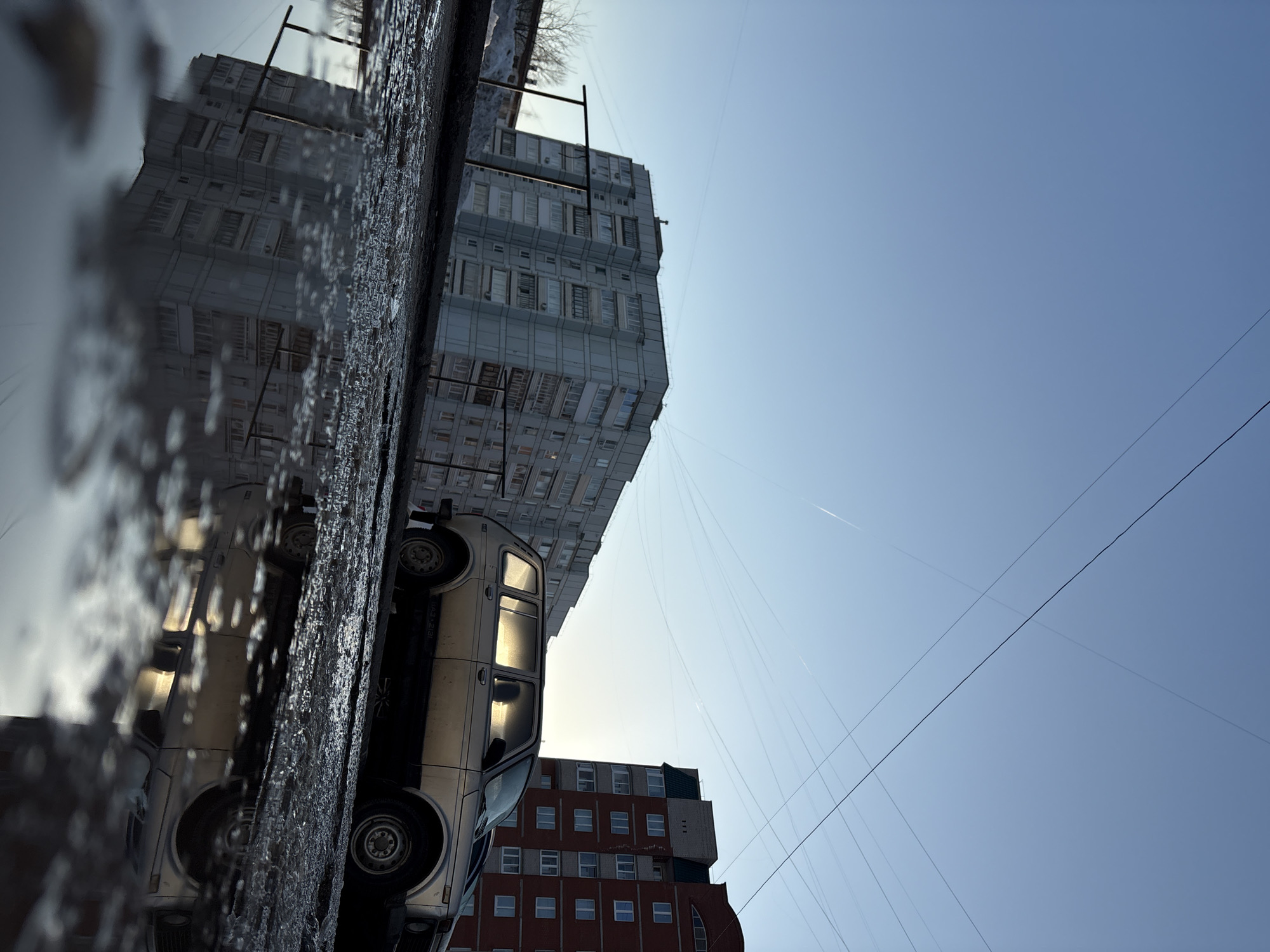 This forensic description details an urban scene in Chaikovsky, Russia. The image is taken from a low perspective, featuring a large puddle dominating the foreground, reflecting the surrounding environment. In the mid-ground, a light-colored Lada Niva (model VAZ-2131, indicated by text on the vehicle's side) is parked on the right. The vehicle's driver-side windows exhibit strong reflections of a low-angle sun, obscuring any interior details or occupants. Behind the Lada Niva, a multi-story reddish-brown brick apartment building with a dark green roof is visible. To the left, a taller, multi-story light grey or white apartment building with numerous balconies is present, its reflection clearly visible in the foreground puddle. The ground around the puddle appears wet, muddy, and potentially icy or slushy, with patches of dirty snow visible to the far left. The sky above is clear and light blue, traversed by multiple thin contrails from aircraft and an intricate network of overhead utility wires. Bare tree branches are discernible on the far left, behind the snow. No individuals or specific actions are depicted.
