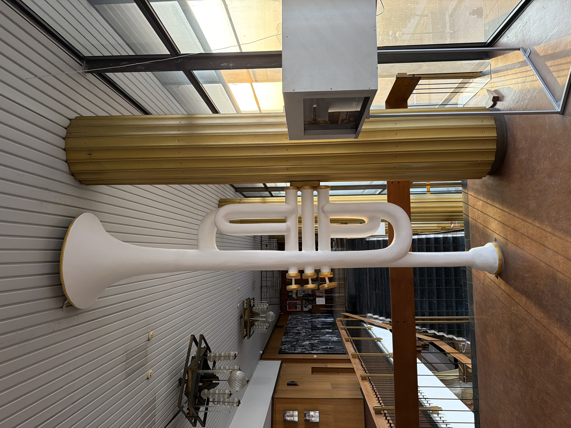 The image depicts the interior of a building, likely a public space or cultural venue, with architectural elements and a large decorative sculpture. Prominently featured is a white, stylized sculpture resembling a brass instrument, possibly a trumpet or tuba, positioned in the foreground. This sculpture is illuminated by natural light streaming from large windows. To the right of the sculpture, a fluted golden column supports the ceiling. A modern display case on a metal stand is also visible near the column and window.
The background reveals a multi-level interior with a wooden railing and a staircase leading upwards. The walls are paneled with wood, and a large black and white mural or collage is mounted on one wall, featuring numerous small portraits or images. Several modern chandeliers hang from the slatted ceiling.
The scene is characterized by warm, natural lighting, casting distinct shadows across the floor and surfaces, suggesting it was taken during daylight hours, possibly late afternoon due to the golden hue of the light. The overall impression is one of a grand, possibly retro-futuristic architectural space. The context provided by the user indicates the location is in Odesa, Ukraine. The specific building is likely a significant cultural institution within the city, given the scale and artistic elements present. There are no people visible in the image.