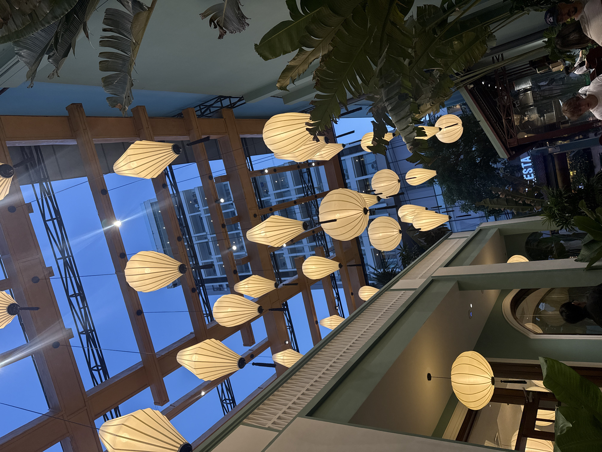 This is a medium shot, angled from a lower perspective, looking upwards at the ceiling of a restaurant in Nha Trang, Vietnam. The ceiling is a combination of a glass roof supported by a wooden beam structure and solid, light green painted architectural elements. Hanging from the ceiling are numerous decorative lanterns of various shapes and sizes, predominantly white and illuminated, creating a warm ambiance. Large, leafy tropical plants are visible in the foreground and on the right side of the frame, adding to the tropical feel. In the background, a modern, glass-fronted building with multiple floors is visible through the glass roof, suggesting an urban setting. The lighting suggests it is dusk or evening. There are several people present in the scene, mostly blurred or partially visible, suggesting they are patrons or staff within the restaurant or on balconies associated with it. One person is looking upwards towards the lanterns. A neon sign that partially reads "ESTA" is visible in the middle ground.
