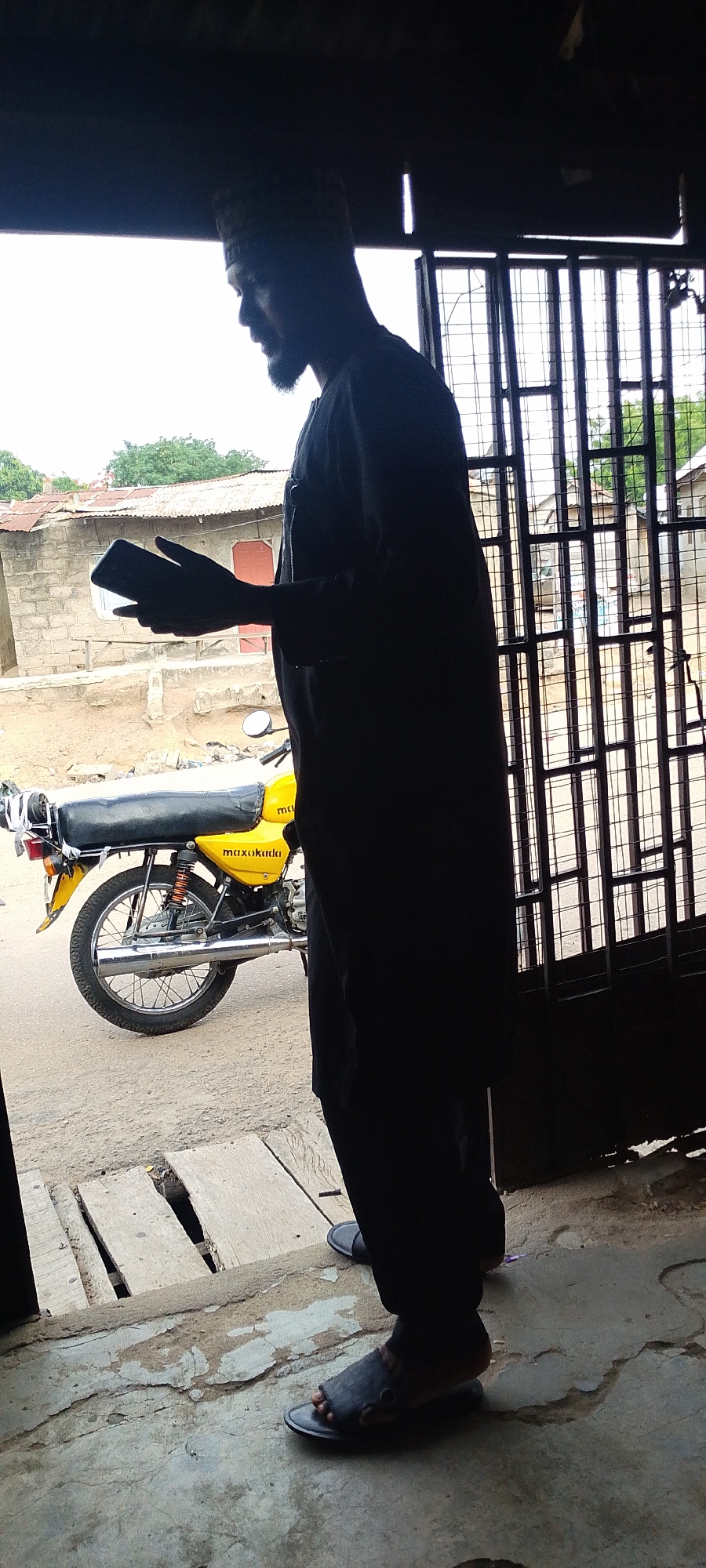 A man is standing in a doorway with a grated metal door at a street in Lokoja, Nigeria. The man is wearing a hat and long robes, and he is holding a mobile phone in his hands. A yellow motorcycle is visible behind him in the street. The environment appears to be a somewhat dilapidated urban area, as can be seen by the street and buildings.