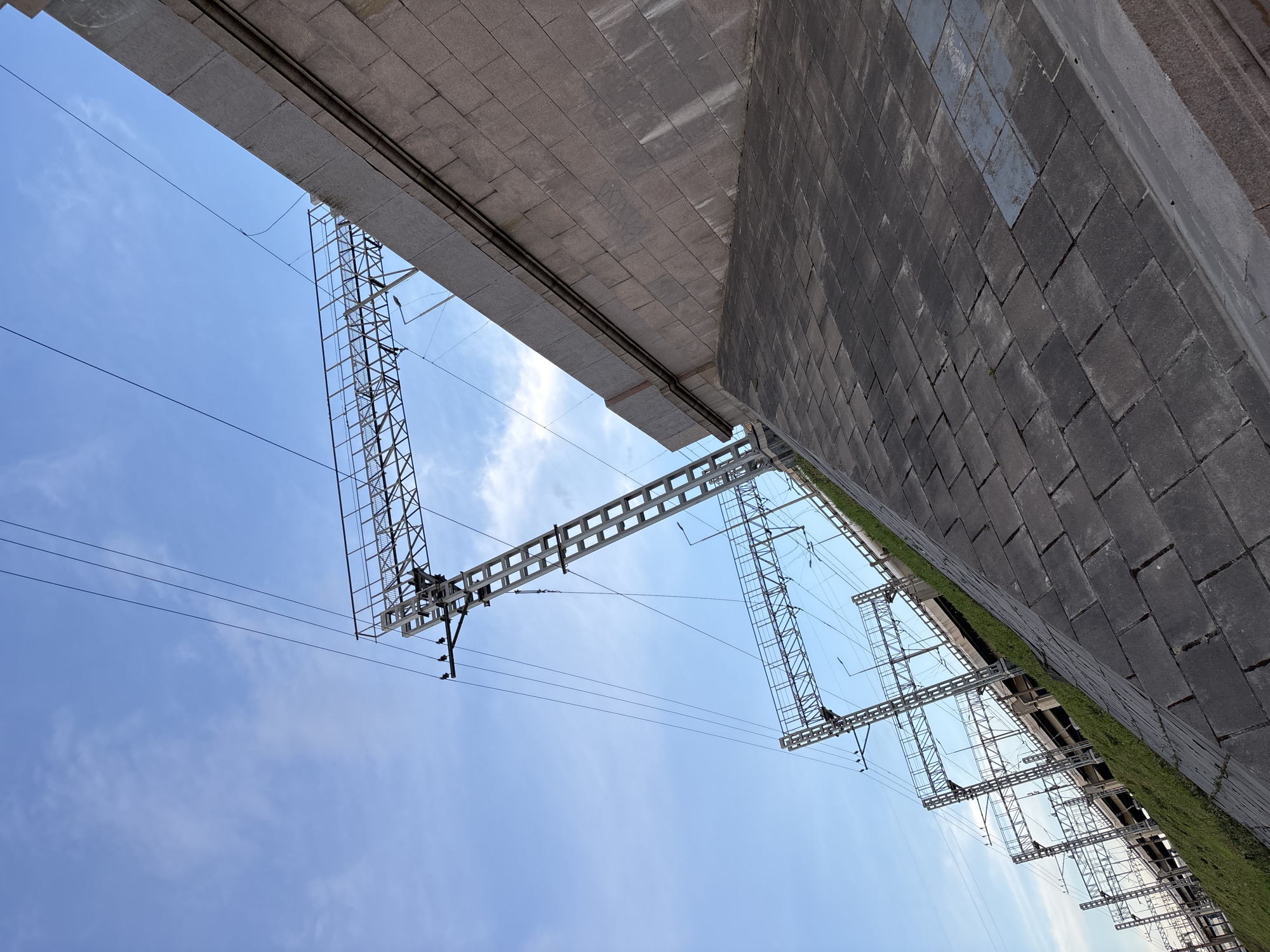 The image displays an upward-looking view of robust infrastructure against a bright blue sky with scattered wispy clouds, suggesting daytime. Dominating the upper left is the underside of a large, grey concrete or stone structure, possibly a bridge or overpass, composed of rectangular blocks. Running parallel to this structure are several overhead electrical lines.
Below this, and extending across the mid-ground, are multiple intricate metal lattice gantries, typical of those used in electrified railway systems or for large-scale power transmission. These gantries feature insulators and support numerous overhead wires, spanning the open sky.
On the right side of the frame, a substantial grey wall, constructed from rectangular masonry blocks, rises steeply. A narrow strip of green grass is visible at the base of this wall where it meets what would be the ground level.
No human activity is visible, and there is no discernible text in the frame. The scene captures a powerful industrial or transportation landscape, consistent with urban infrastructure found in places like Saint Petersburg, Russia.