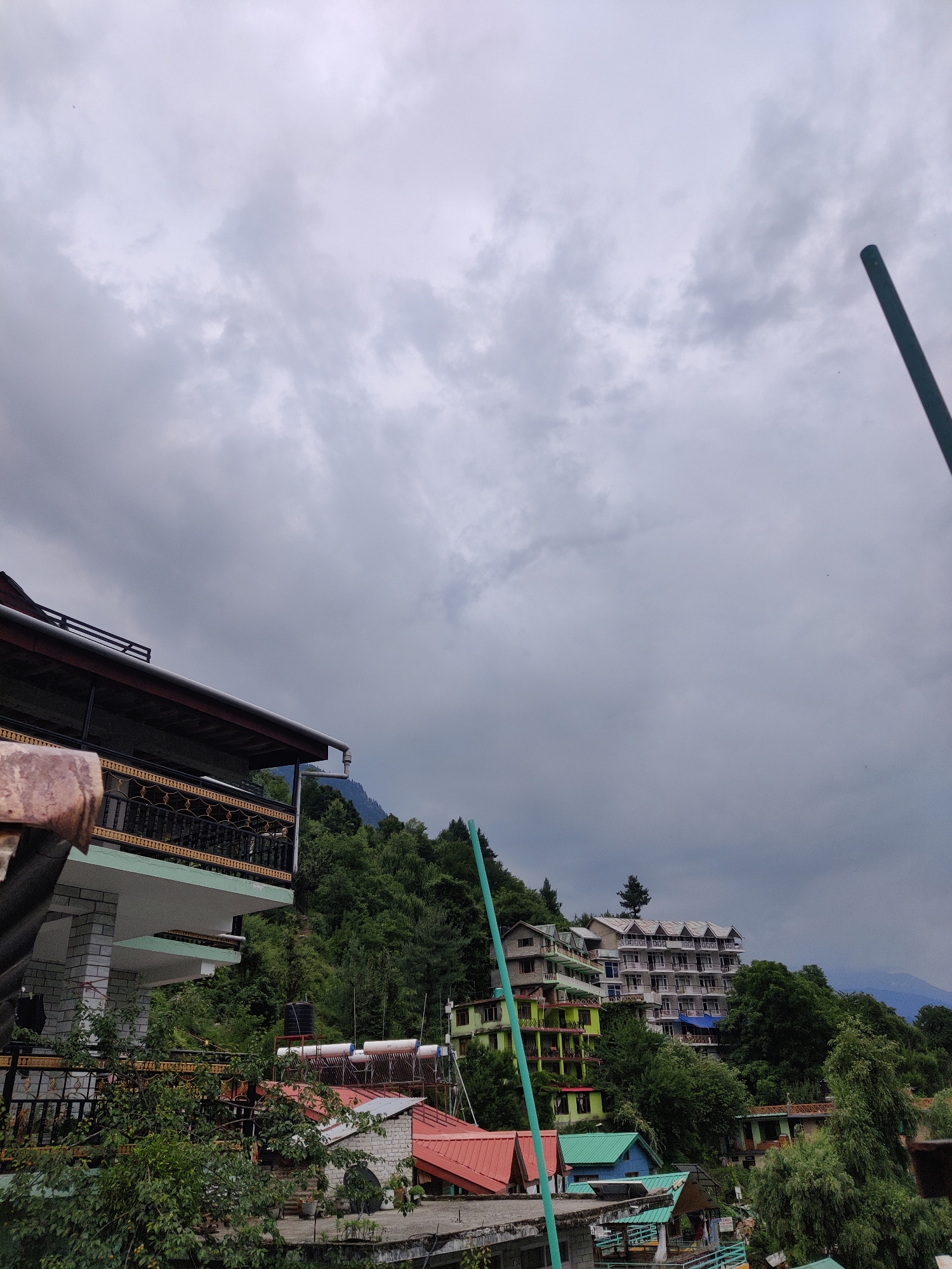 The image depicts a cloudy day in Vashisht, India, showcasing a hillside covered with lush green trees and several buildings. In the foreground, a building with a balcony featuring ornate railings is visible. Further up the hill, a multi-story building with a white facade and many windows stands prominently. Below this, other structures with colorful roofs, including a bright green one, are interspersed with the dense foliage. A tall, thin green pole or antenna extends vertically into the frame from the lower left. In the distance, a range of mountains is faintly discernible through the overcast sky. The scene suggests a residential or tourist area built into the natural landscape.