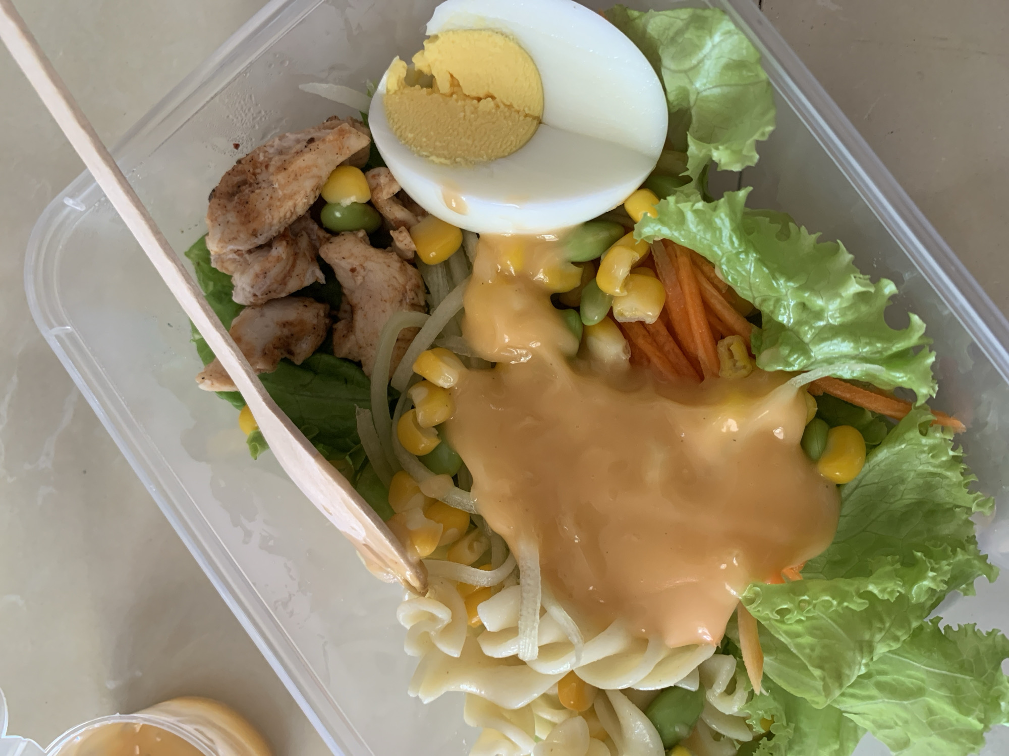 The image shows a close-up, overhead view of a salad in a clear plastic container. The salad consists of rotini pasta, corn, edamame, shredded carrots, lettuce, sliced hard-boiled egg, and grilled chicken pieces. A wooden stick is positioned diagonally across the top of the salad, and a creamy orange dressing is poured over a portion of the ingredients. The container is sitting on a light-colored, textured surface, possibly a table. A small, partially visible jar with a yellow liquid is present in the upper left corner. The lighting is bright, suggesting an indoor setting. There are no people visible in the image. The image does not provide any information or visual cues that can be used to determine its location in Sukawati, Indonesia.