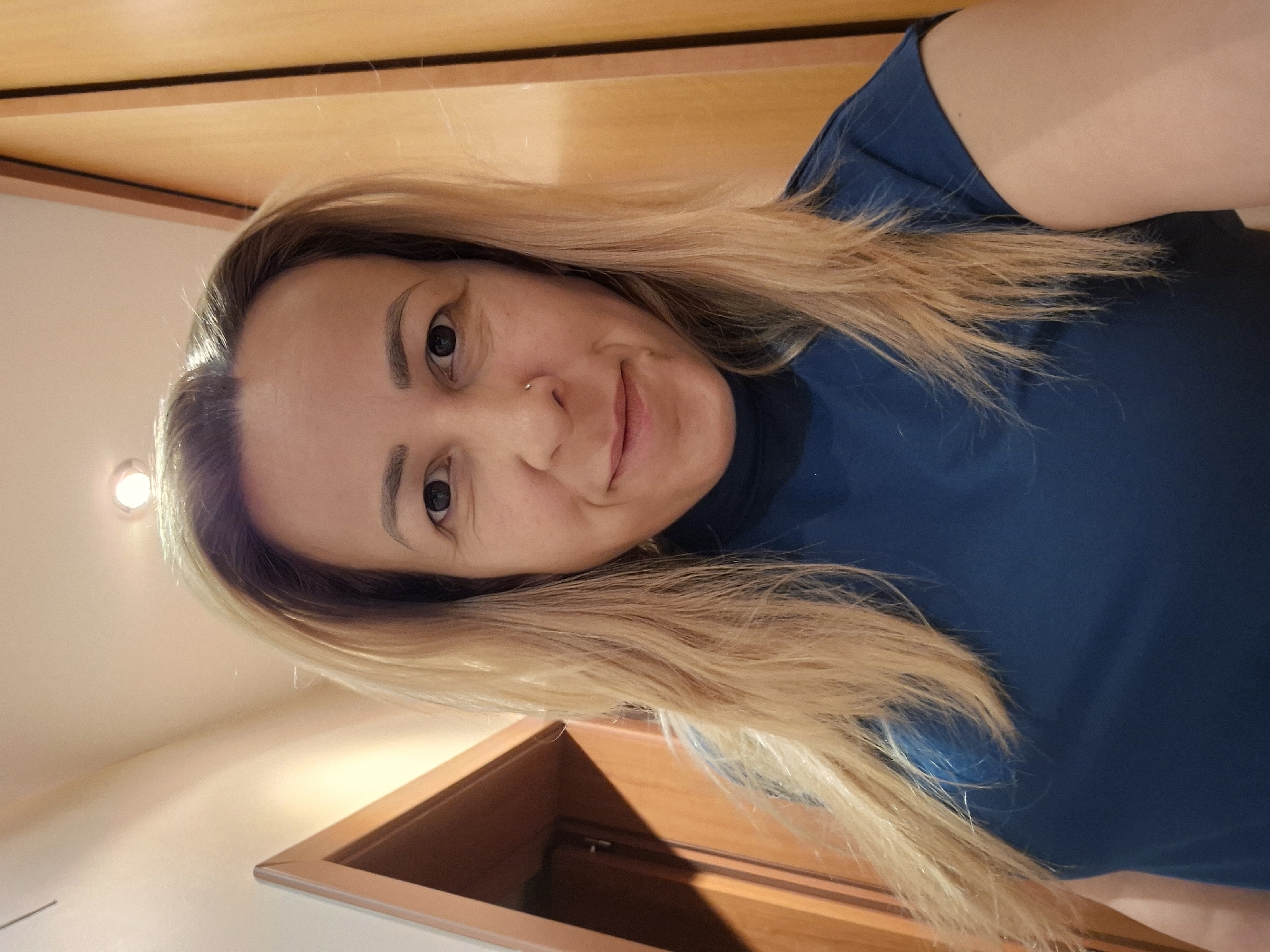 A woman with blonde hair and a nose ring is posing for a selfie indoors. She is wearing a blue shirt and standing in front of a light-colored wooden door or wall. The location is Unknown, Unknown.