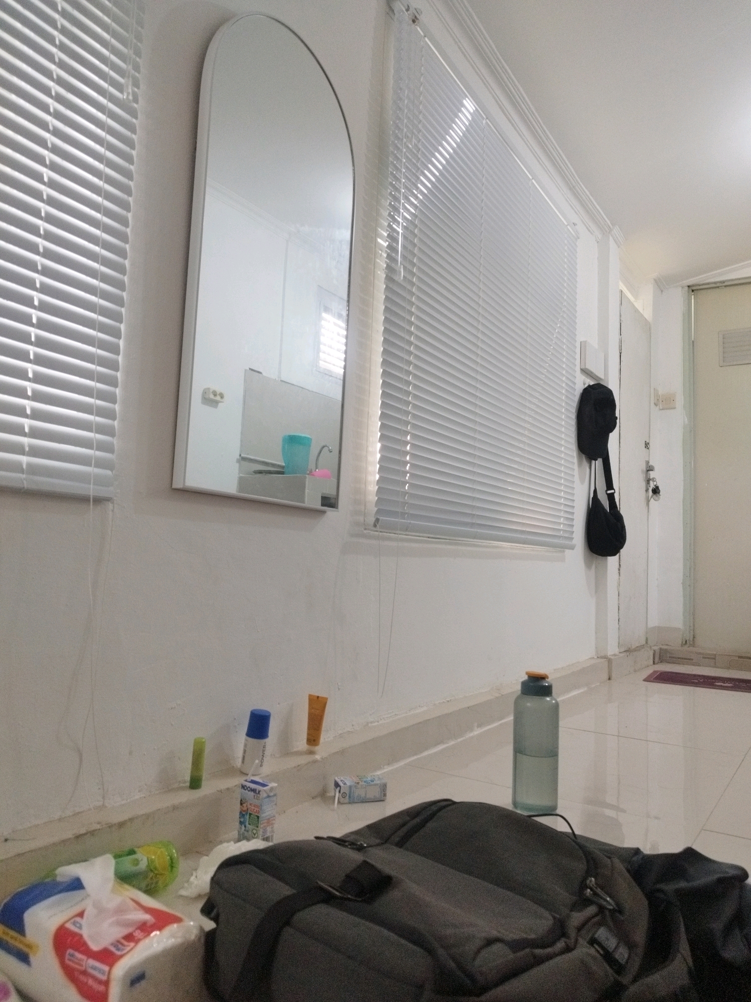 The image depicts an indoor scene, likely a residential hallway or room, with a focus on a wall adorned with blinds and a mirror. The overall lighting suggests daytime, with natural light filtering through the blinds. The floor is tiled and appears clean.
On the wall to the left, there is a large, arched mirror reflecting a portion of what appears to be a bathroom or kitchenette area, showing a white sink, a blue cup, and a faucet. To the left of the mirror, a window is covered by white horizontal blinds.
Along the base of the wall, a narrow ledge or shelf holds several small objects, including a green tube, a blue-capped bottle, a small orange tube, a carton of milk, and another small box.
In the foreground, a dark-colored backpack rests on the floor. Next to it, to the left, is a package of facial tissues. A clear plastic water bottle with a light-colored cap is positioned on the floor to the right of the backpack, partially filled with a clear liquid.
On the right side of the image, a doorway is visible, leading to another area that is out of focus. Two black bags are hanging on a hook on the wall near the doorway. The scene is tidy, with no obvious signs of disarray, suggesting a well-maintained living space.
There are no individuals present in the frame. The image is a still photograph of an interior environment.
The scene does not contain any elements that would specifically identify it as being located in Sukabumi, Indonesia, or any other specific city or country. The objects and furnishings are common to many residential interiors globally.