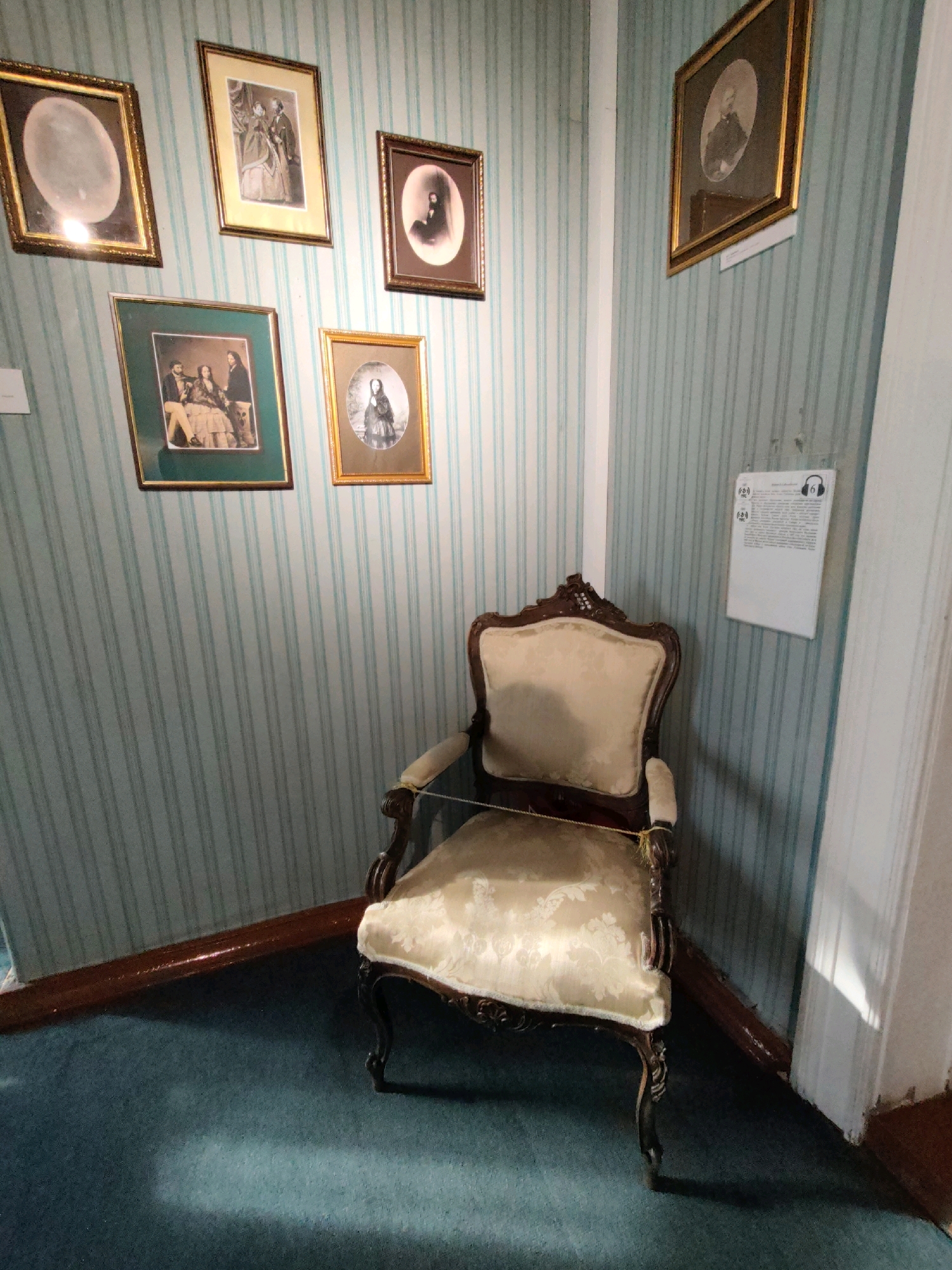 The image depicts a corner of a room with teal and white striped wallpaper and a dark blue carpet. A single ornate armchair with light-colored patterned upholstery and dark wooden legs is positioned in the center of the frame. A thin yellow cord is tied around the arms of the chair. On the walls are several framed photographs, mostly portraits of individuals in historical attire. Some of the frames are gold, and others appear to be darker wood. There is also a white informational plaque with some text and icons hanging on the wall to the right of the chair. The lighting suggests an indoor setting, possibly a museum or historical display, given the arrangement of the furniture and framed items. This scene is located in the Volkonsky House Museum (Dom Volkonskogo) in Irkutsk, Russia, a preserved estate associated with the Decembrist exiles.