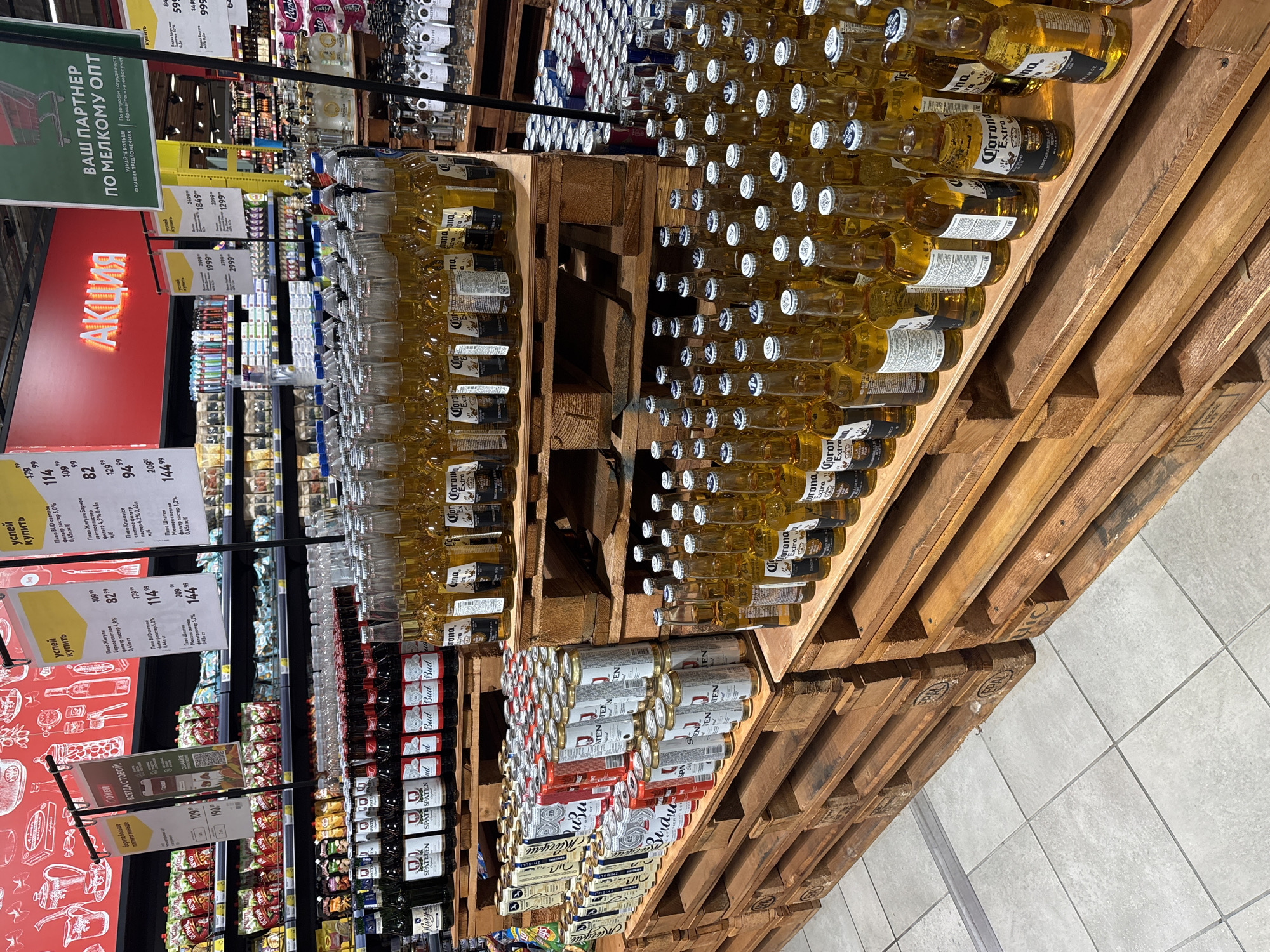 The image shows a display of alcoholic beverages in what appears to be a supermarket or retail store in Sochi, Russia. Pallets are stacked with Corona beer bottles and beer cans. Various other beverages are visible on the shelves in the background. Price tags and promotional signage are visible in Russian, indicating sales and bulk purchase options. The floor is tiled in light gray.