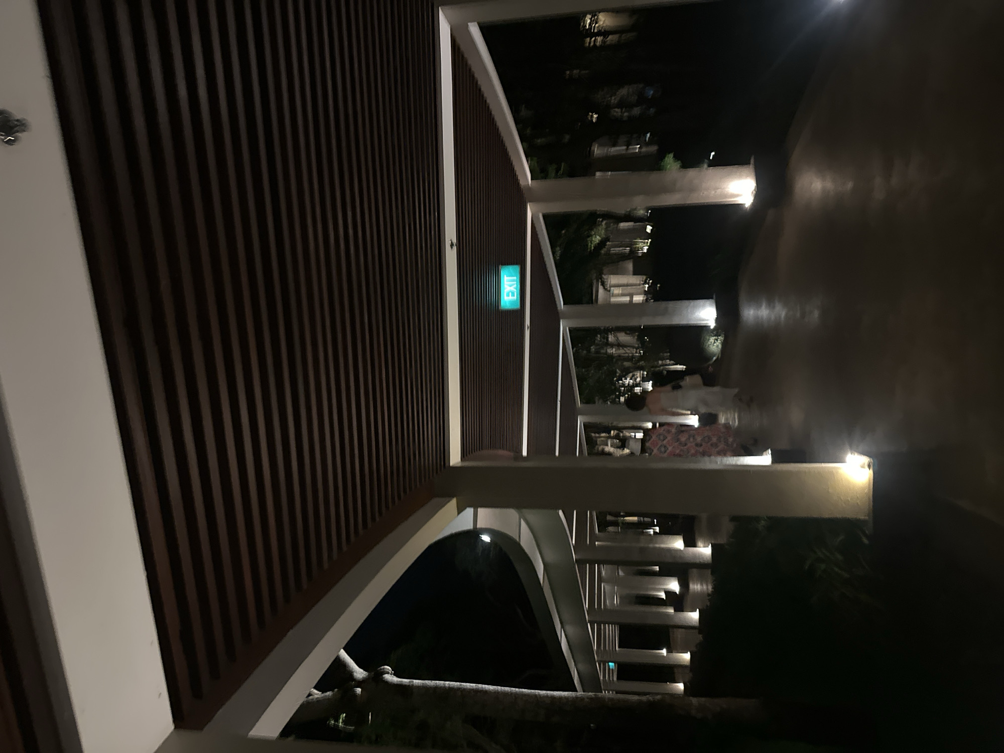 The image displays a modern, illuminated covered walkway at night, likely part of a resort or architectural complex in Kuta Selatan, Indonesia. The perspective looks along the pathway, which curves slightly to the right.
To the left, a prominent wall or ceiling section is clad with dark, vertically oriented wooden slats. A bright green "EXIT" sign is clearly visible against this slatted surface. The pathway is bordered by a series of white structural beams and columns that form arches, extending into the distance and creating a sense of depth. Small, warm-toned light fixtures are integrated into these white columns, casting illumination onto the pathway and reflecting gently on its surface.
In the mid-ground on the right side of the walkway, two blurry figures, likely people, are seen walking away from the viewer. One figure appears to be wearing a patterned dress. Beyond the illuminated structures, the background is dark, with vague hints of trees or foliage. Beneath one of the archways on the left, a distinct, light-colored, gnarled tree trunk is visible. The overall scene conveys a tranquil, well-lit, and architecturally refined outdoor space at night.
