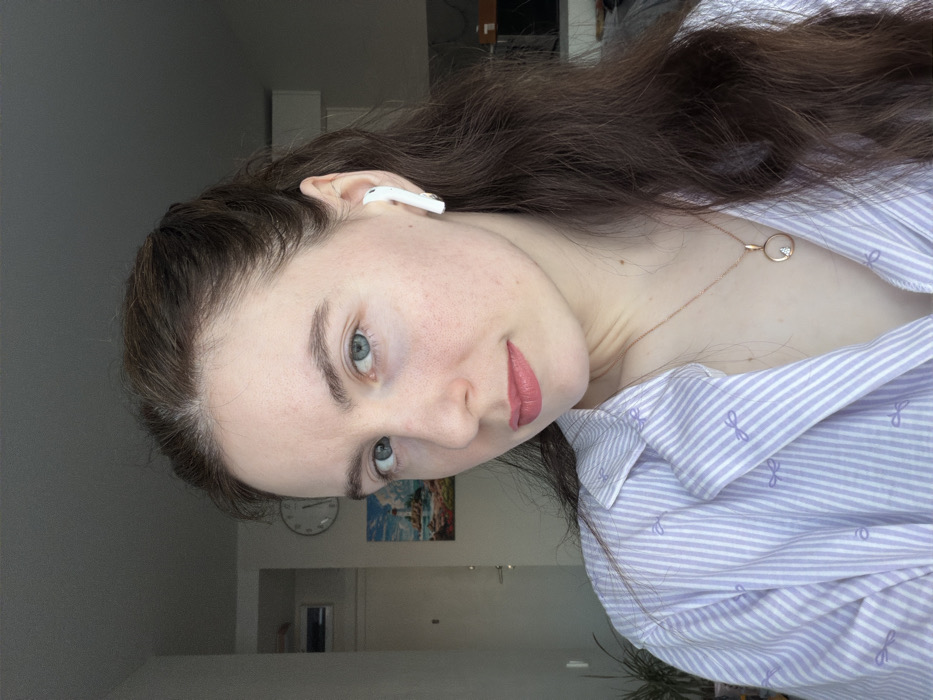 The image displays a headshot and upper torso of a female individual in an indoor residential setting within the city of Ковалёво, Russia. The subject, possessing fair skin, long dark brown hair, and light blue/green eyes, gazes directly at the camera with a neutral expression. She wears a light purple and white striped collared shirt featuring small bow patterns and a rose gold-colored necklace with a circular pendant containing a clear gemstone. A white wireless earbud is present in her right ear. The background shows a light-colored wall with a partially visible door on the left, an overhead rectangular vent, a circular wall clock, and a framed picture depicting a coastal scene with a lighthouse. A portion of a green potted plant is visible in the lower left foreground. The background to the right is darker and less defined.