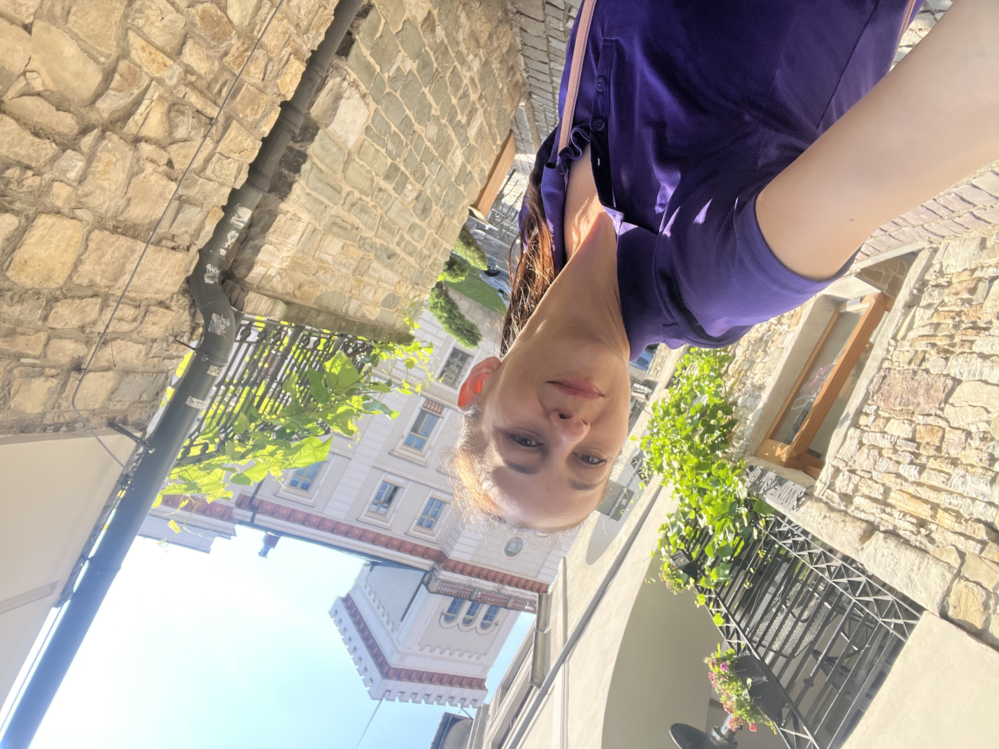A young woman is taking a selfie in an outdoor European-style alleyway. She is wearing a purple t-shirt and has her hair down. Her face is partially visible, and she is looking at the camera with a neutral expression. The alleyway is lined with stone buildings and has greenery growing on some of the walls. There is a tower visible in the background, and the sky is clear and blue, indicating a sunny day. The lighting suggests it is daytime, possibly late morning or early afternoon. A sign on the building reads "WINE SHOP". The overall scene depicts a casual moment in a picturesque urban environment.