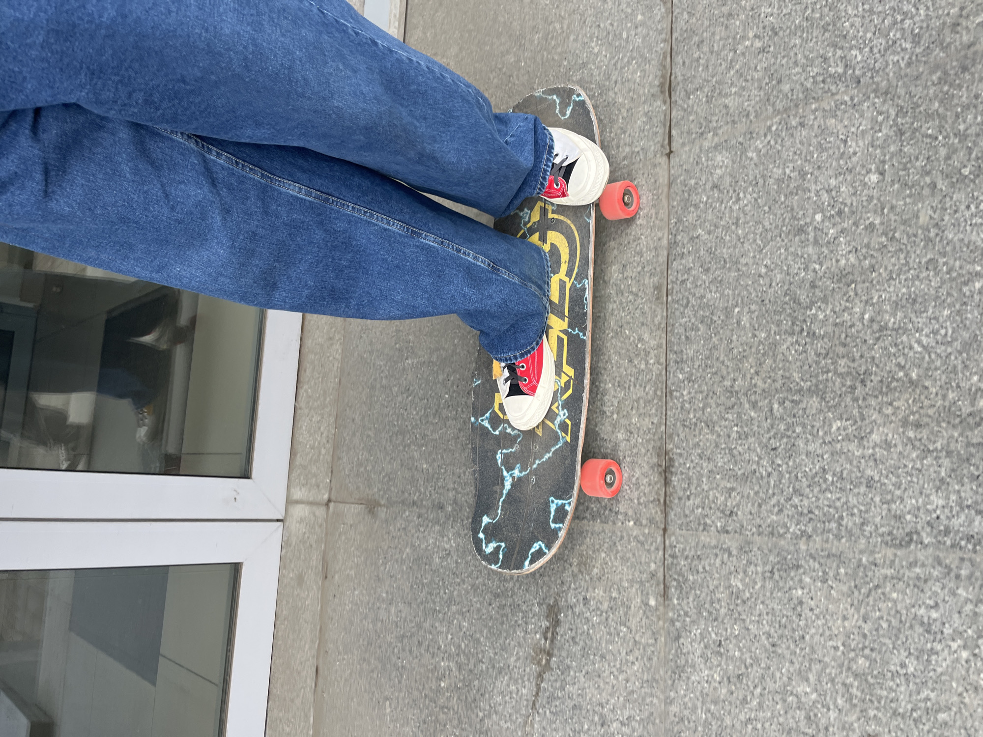 A person wearing denim jeans and white and red shoes is standing on a skateboard on a gray tiled surface. The skateboard has a black base with a yellow logo and blue lightning bolt designs. The wheels of the skateboard are orange. To the left of the person are white doors/windows. The location appears to be outdoors.