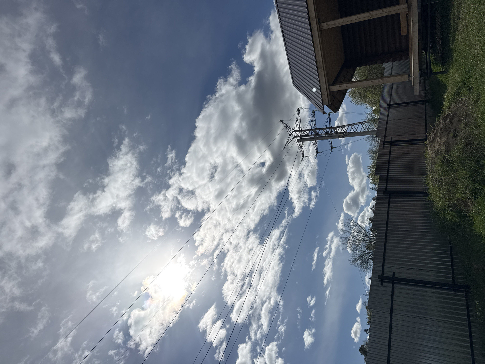 The image shows a low-angle view of a cloudy sky with an electrical transmission tower in the city of Chaikovsky, Russia. Electrical wires extend from the tower towards the lower left of the frame, where the sun shines through. To the upper right of the tower, a portion of a building's roof with exposed rafters is visible. A corrugated metal fence runs along the right side of the image, with grass and some vegetation visible beyond it.