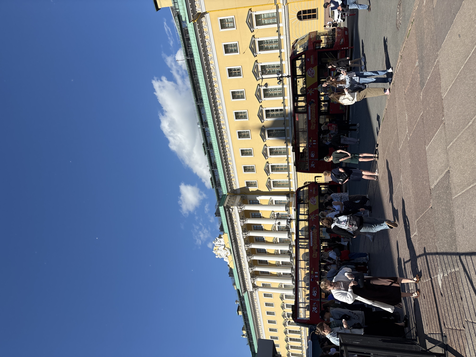 The image depicts a sunny day with a clear blue sky and scattered clouds in Saint Petersburg, Russia. In the foreground, a group of people are standing on a paved area, some looking at their phones. Behind them, a bright yellow, multi-story building with a green roof and classical columns is visible. Red double-decker buses with "City Sightseeing" written on them are parked in front of the building, suggesting a tourist area. The building appears to be a historical landmark, possibly a palace or government building, given its grand architecture. The presence of tourists and sightseeing buses indicates that this is a popular attraction within the city.