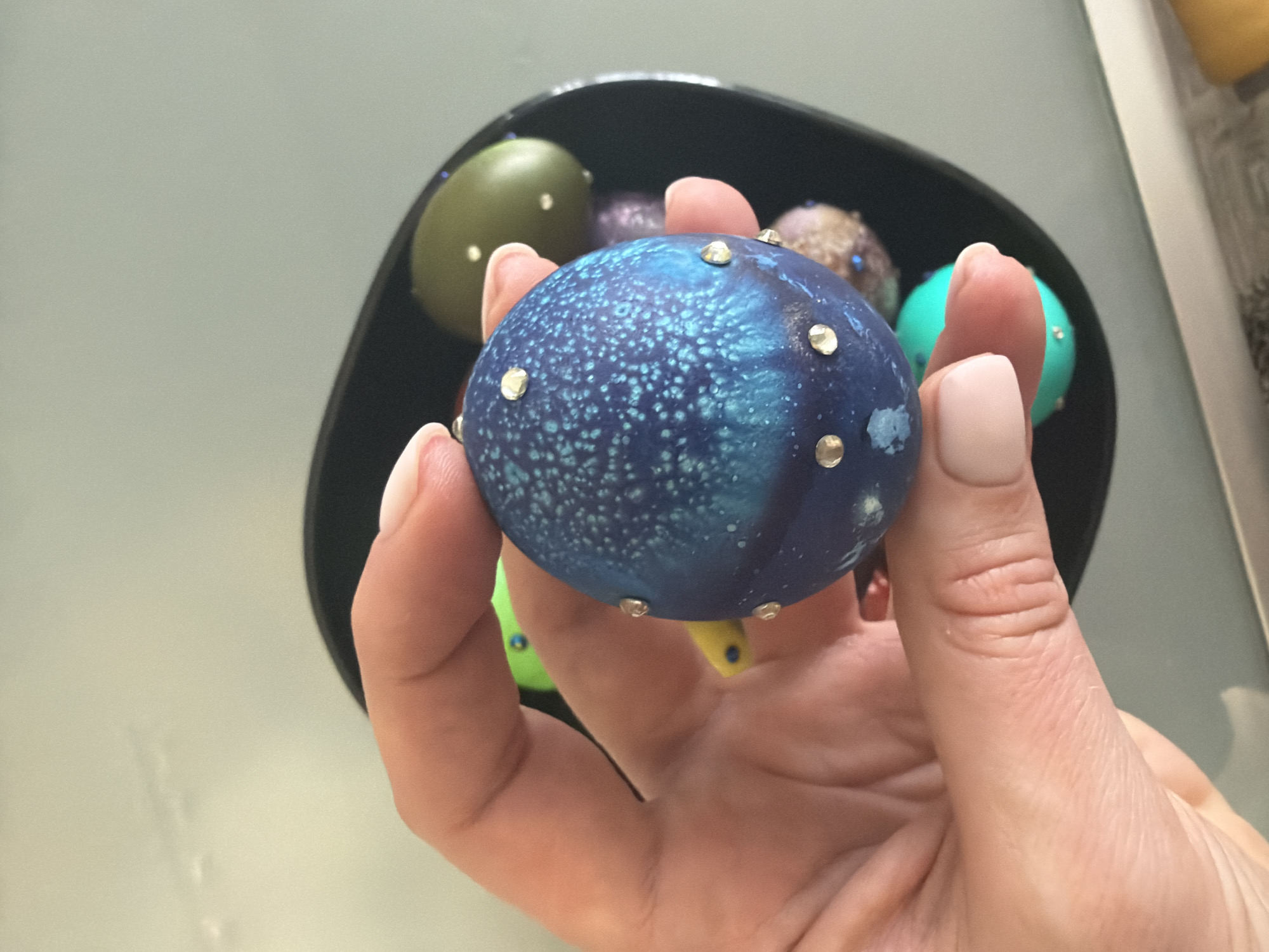 A hand holds a blue, speckled, ornamented egg in front of a black bowl filled with similarly decorated eggs in various colors. The background is out of focus, revealing a gray surface and a white object. No evidence suggests this scene is in Moscow, Russia.