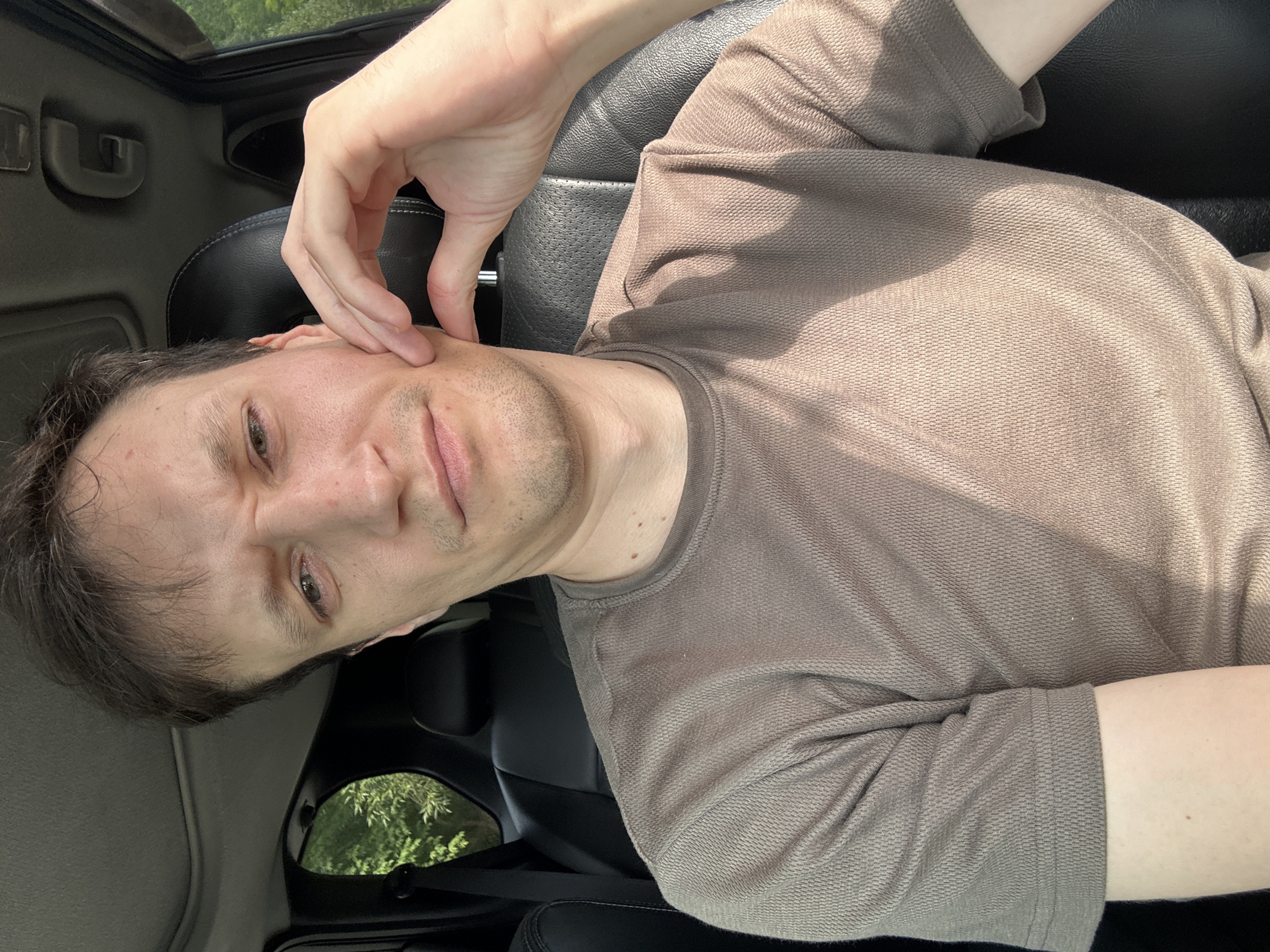 A young Caucasian man is sitting in the driver's seat of a car, with his left hand resting on his cheek. He is looking directly at the camera with a slightly furrowed brow. The interior of the car is visible, including black leather seats and a sunroof. Outside the car window, trees and greenery can be seen, suggesting a rural or suburban setting. The image appears to have been taken during the daytime, with sunlight casting shadows on the man's face and shirt. There is no information available to confirm the location within Makiv, Ukraine.