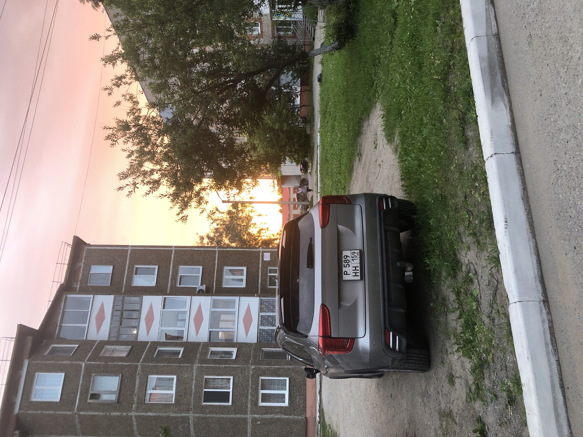 This media file captures an exterior view of a residential building and a parked car in Solikamsk, Russia. The scene is dominated by a multi-story building with a concrete facade, featuring numerous windows and distinct diamond-shaped decorative elements on some levels. A gray car is parked on a dirt path next to the building, facing away from the viewer. The car's license plate is visible, displaying "P 589 HH 159" and "RUS". A curb and a paved area are also present in the foreground. Lush green grass grows along the side of the path and near the building. In the background, trees with dense foliage are visible, with a building structure peeking through. The sky is awash with the soft hues of sunset, casting a warm glow over the scene, with shades of pink and orange dominating. The overall atmosphere suggests a peaceful evening in a residential area.