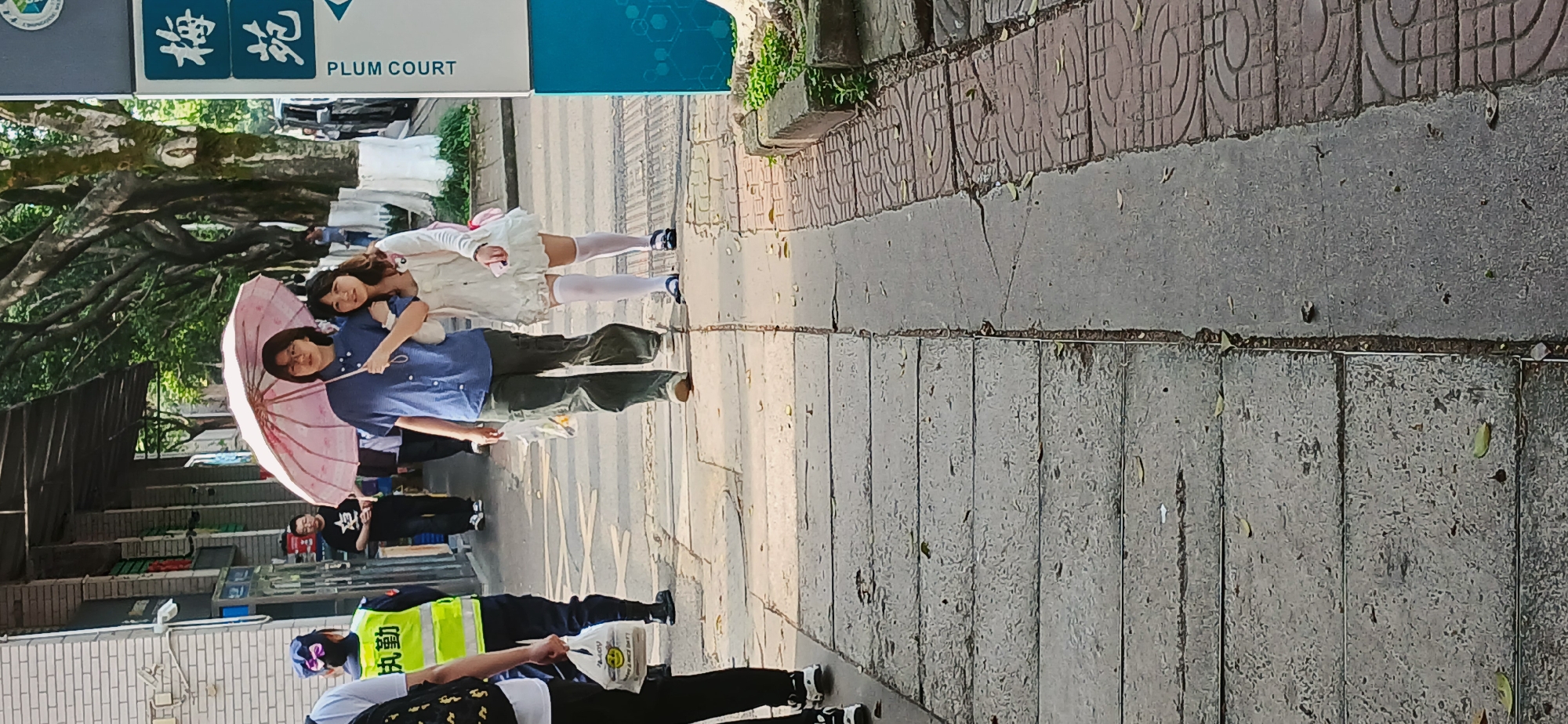 Two young women are on a sidewalk, the woman in the front is carrying a pink umbrella. The woman in the back is wearing a white dress. They are under a tree near a sign with “PLUM COURT” on it. Behind them on the sidewalk are three people, one of whom is wearing a bright yellow vest and carrying a white plastic bag. The image is rotated counterclockwise by ninety degrees. The location is Chongqing High-tech Zone, China. The sidewalk is made of rectangular stones of different colors and patterns.