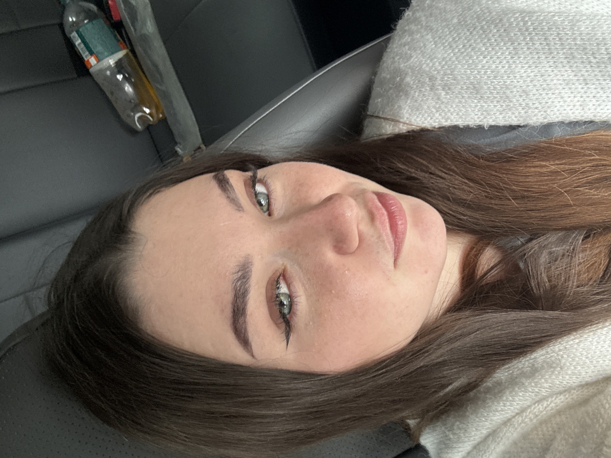 A close-up, angled selfie of a woman with fair skin, gray-green eyes, and dark brown hair. She is wearing a light-colored, possibly knitted, sweater. In the background, partially visible, is the interior of what appears to be a car. A water bottle, partially consumed, is also visible. There is no information to suggest the location is Perm, Russia.