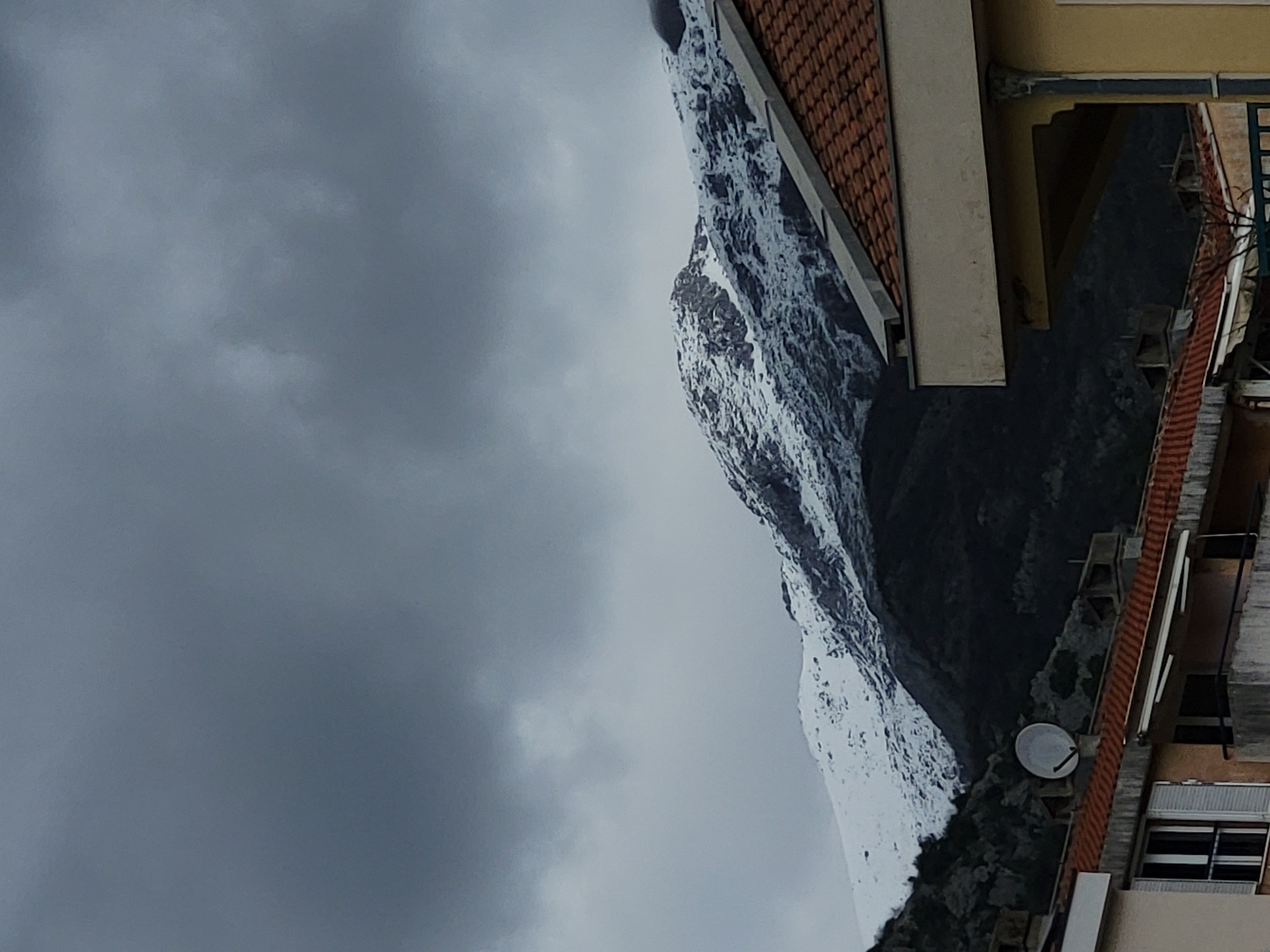 The image presents a static, wide-angle view of snow-covered mountains towering over residential structures in Budva, Montenegro, under a heavily overcast sky. The upper two-thirds of the frame are occupied by a dense, uniform grey cloud layer, indicative of cloudy weather. Below the sky, a rugged mountain range dominates the background, with its higher elevations extensively covered in white snow, revealing patches of dark, exposed rock and sparse vegetation at lower altitudes.
In the midground and foreground, parts of several residential buildings are visible. On the right, a section of a building features a light beige or yellow facade with a reddish-brown tiled roof, and a decorative column-like element on its corner. To the left, a segment of another building shows a similar reddish-brown tiled roof. A white, circular satellite dish is mounted on this roof, positioned against the darker, lower slopes of the mountain. Below the satellite dish, a window with partially open white louvered shutters and a dark metal security grille is visible. No individuals are present, nor are any actions or interactions depicted.