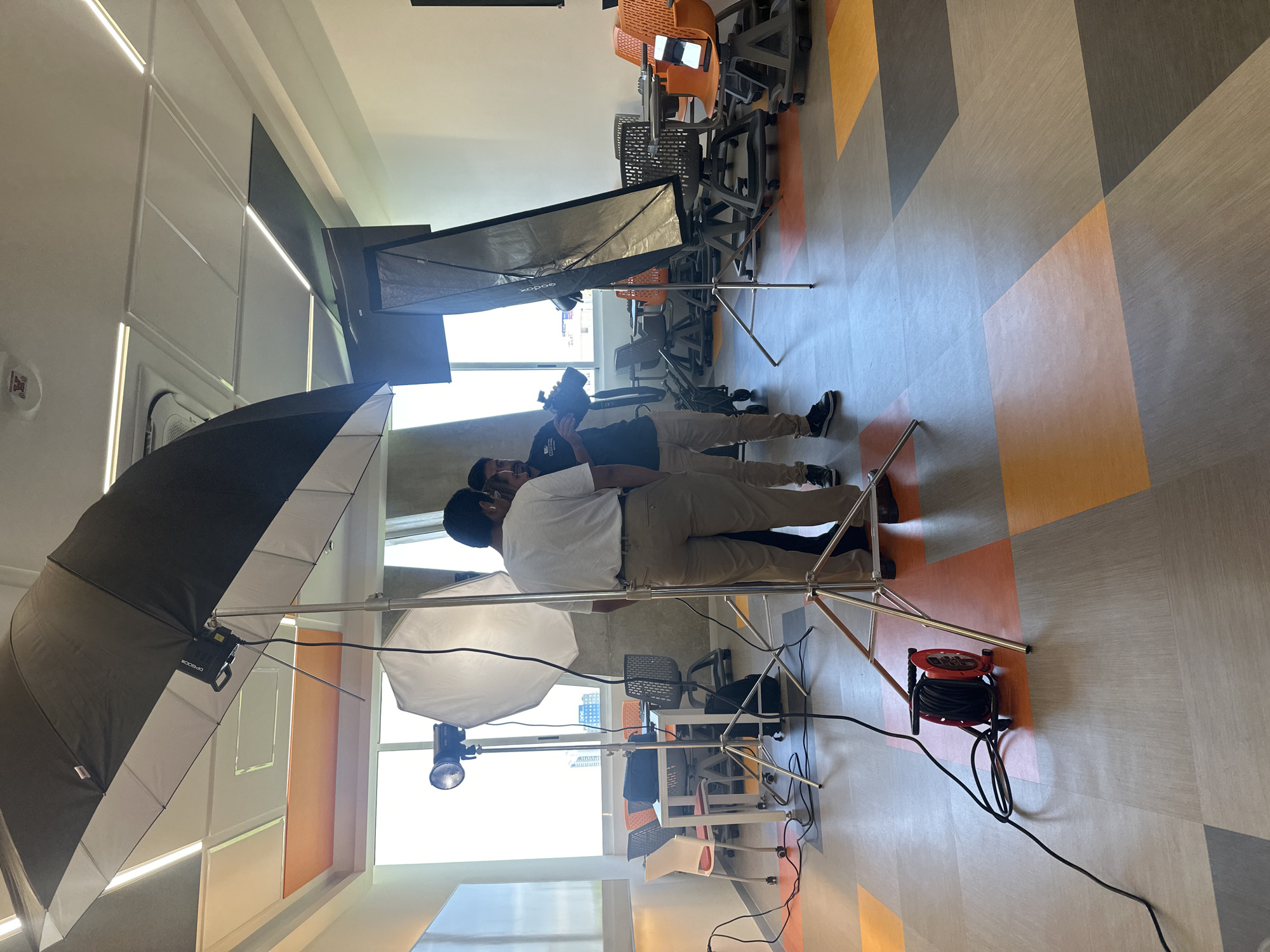The image depicts a behind-the-scenes setup of a photo or video shoot in what appears to be an office space. Two people are present: one is operating a camera, while the other stands in front, likely the subject of the shoot. There are several lighting equipment pieces, including a large umbrella reflector, a softbox, and another smaller light source. Cables and wires are running across the floor, connected to the lighting equipment. In the background, there are office chairs, tables, and windows, offering a glimpse of the exterior. The floor has a patterned design with geometric shapes in varying shades of grey, orange, and beige. A cable reel rests on the floor. The location is not verifiable as San Bernardino Tlaxcalancingo, Mexico, based on the image alone.