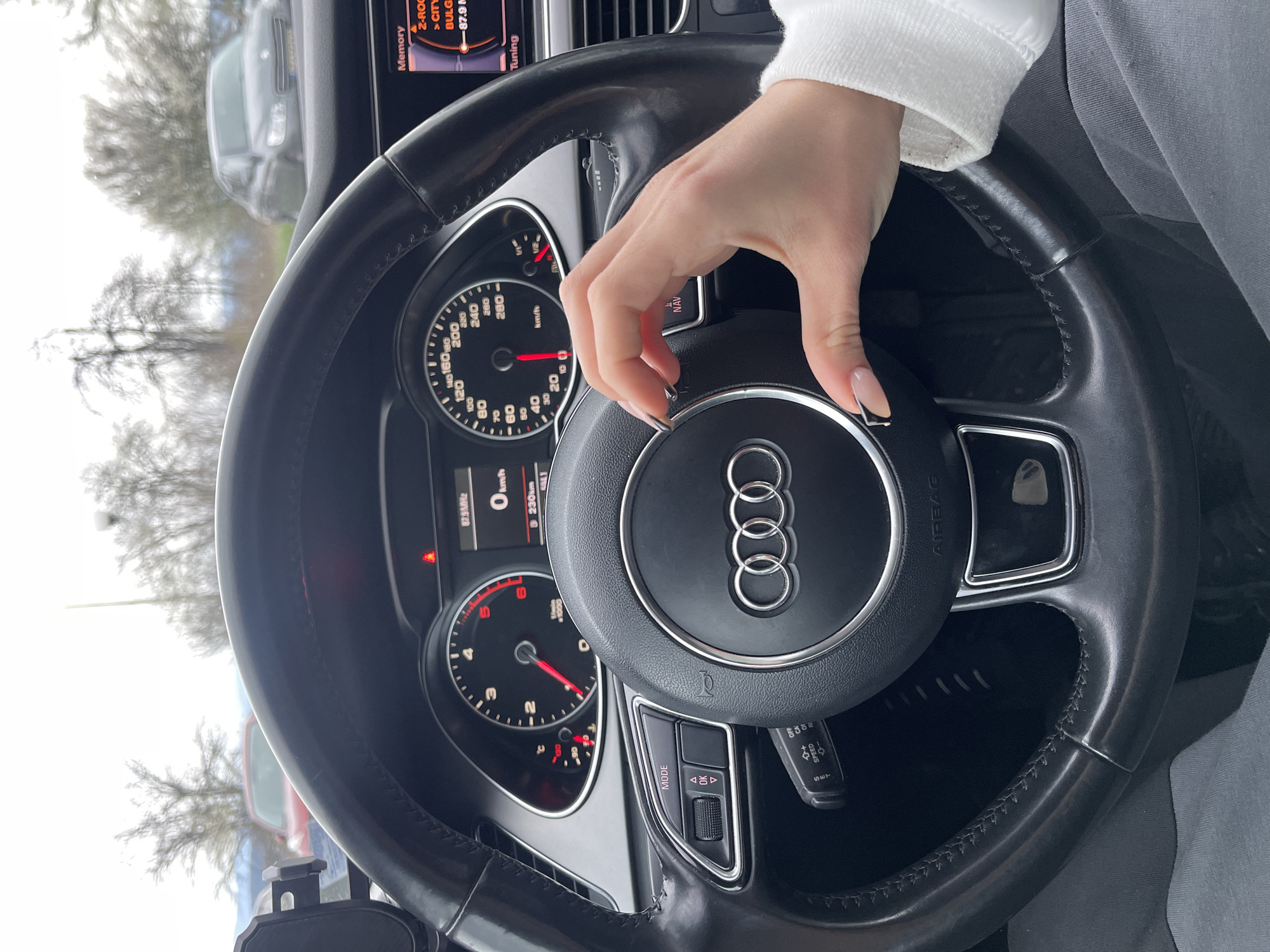 The image shows the interior of an Audi car. The focus is on the steering wheel, featuring the Audi logo in the center. A hand with manicured nails is resting on the steering wheel's central hub. The car's dashboard is visible, displaying gauges for speed, engine RPM, and fuel level. A digital display panel to the right shows radio frequencies, likely indicating the driver is parked somewhere in Bulgaria, in the city of Veliko Tarnovo. Through the windshield, trees and other cars are vaguely visible, suggesting the car is parked outdoors in a lot or street.