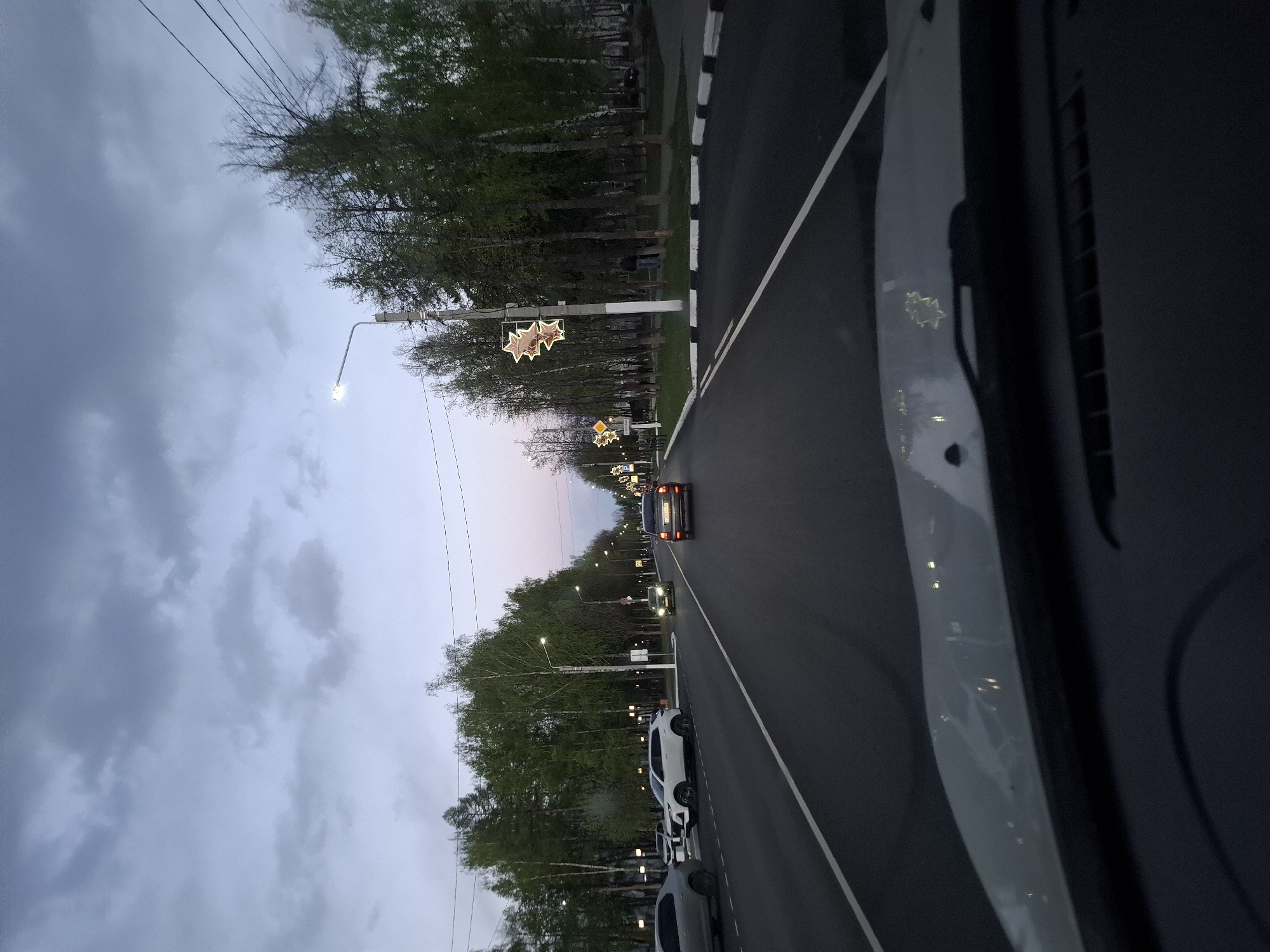 The image depicts a street in the city of Chaikovsky, Russia at dusk. The view is from inside a vehicle looking down the street. The sky is overcast. Street lights are on, decorated with star-shaped ornaments. Cars are driving on the street. Parked cars line the right side of the road. Trees are on both sides of the street. A white line divides the road into lanes. A black and white striped curb is on the right side of the road.