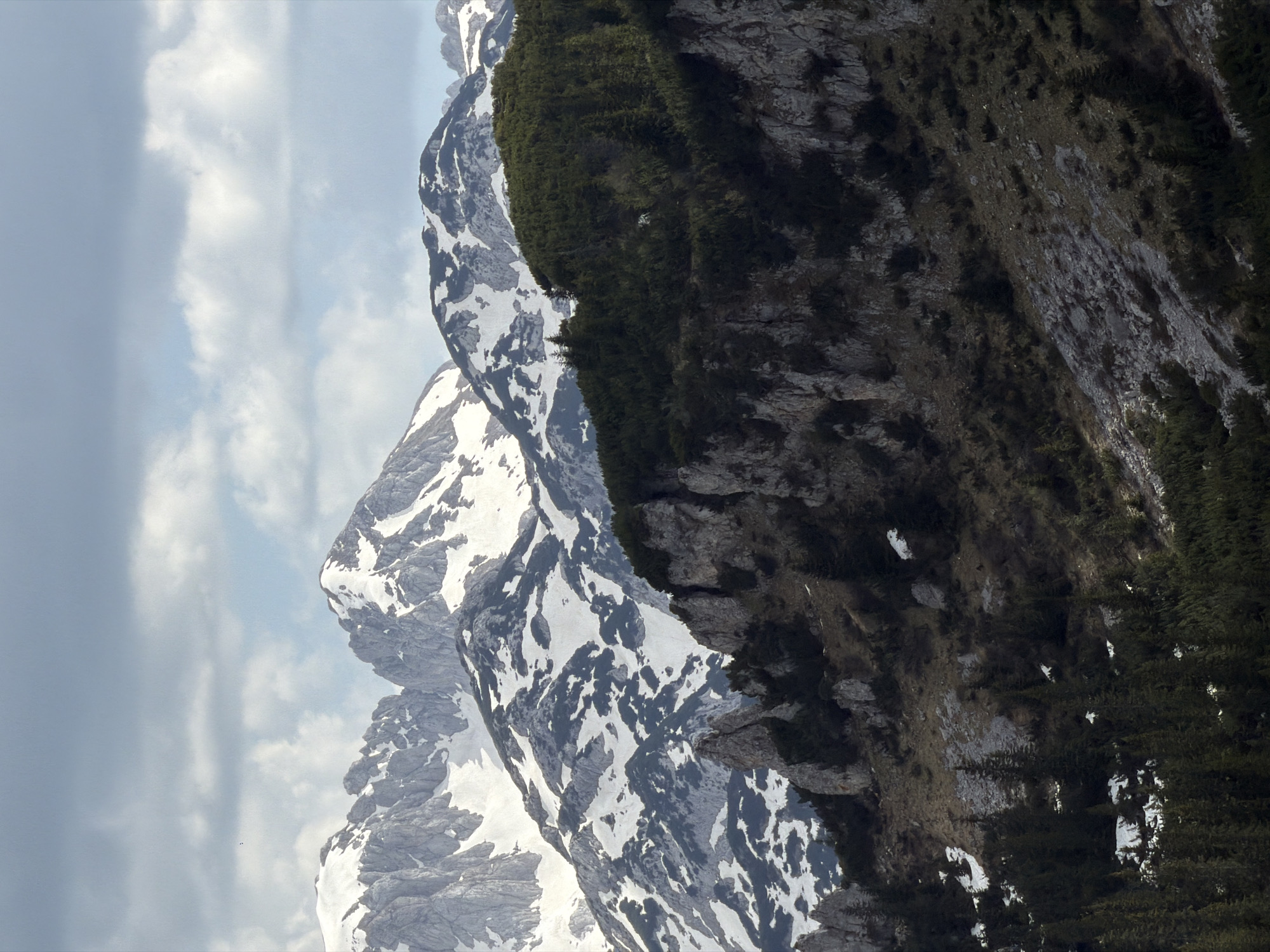 The image shows a mountain range in Bosača, Montenegro. The leftmost peak is partially covered in snow and surrounded by a cloudy sky. The middle and rightmost peaks show bare rock and greenery.