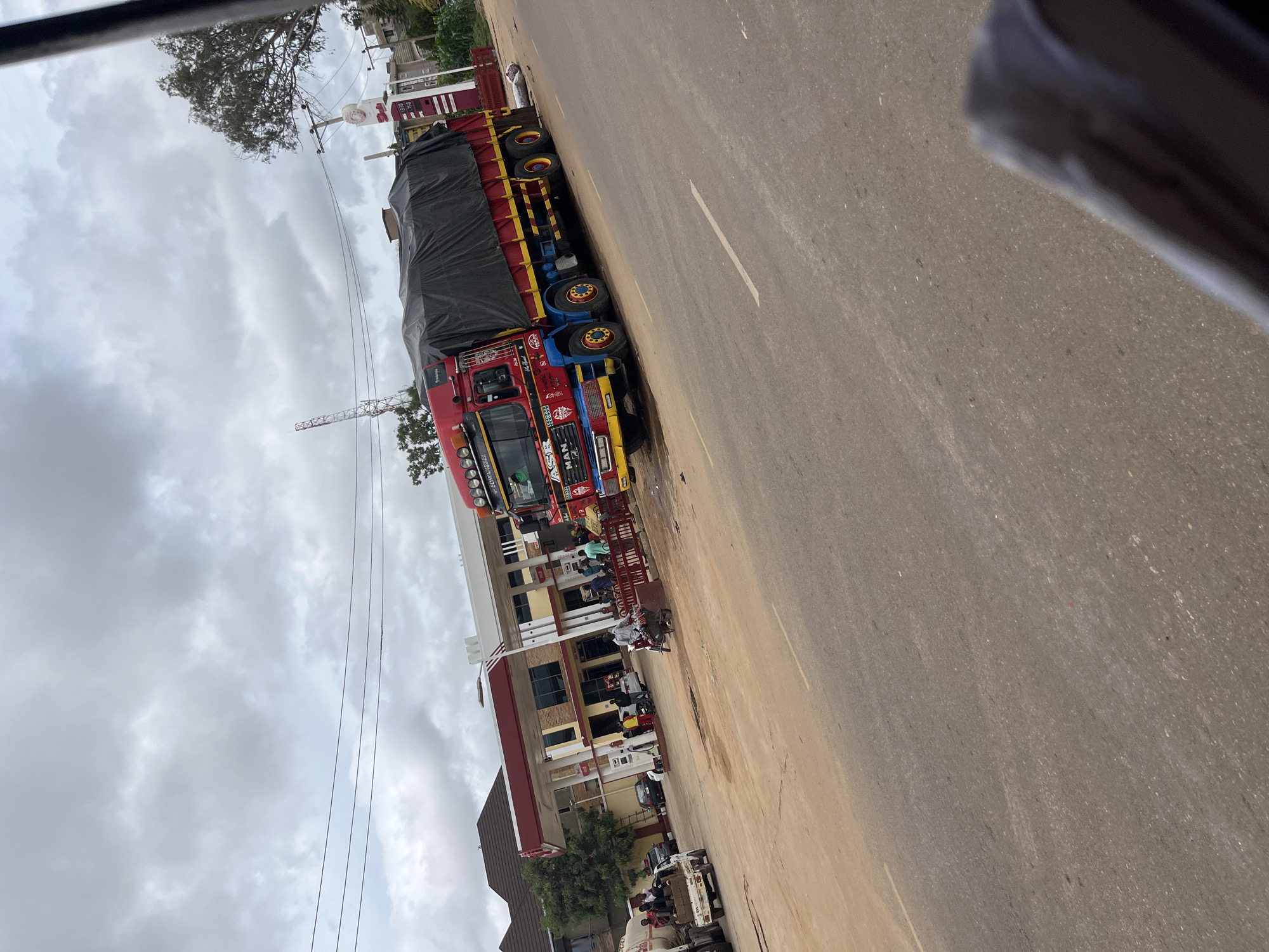 In the city of Rafin Par, Nigeria, a busy street scene unfolds in front of a modern gas station. The sky is overcast, with heavy clouds casting a diffused light over the area. A large, red and blue semi-truck, covered with a black tarp, dominates the right side of the image. It's parked on the road, its colorful decorations and multiple wheels a prominent feature.
To the left of the truck, the gas station building is visible with its red and white canopy. Several cars are parked at the pumps, and people are milling around, some appearing to be refueling or interacting. A smaller white truck with several people sitting in its cargo bed is also present near the gas station, along with a motorcycle. The road surface appears to be asphalt, with painted lane markings. The general environment suggests an active commercial area within the city.