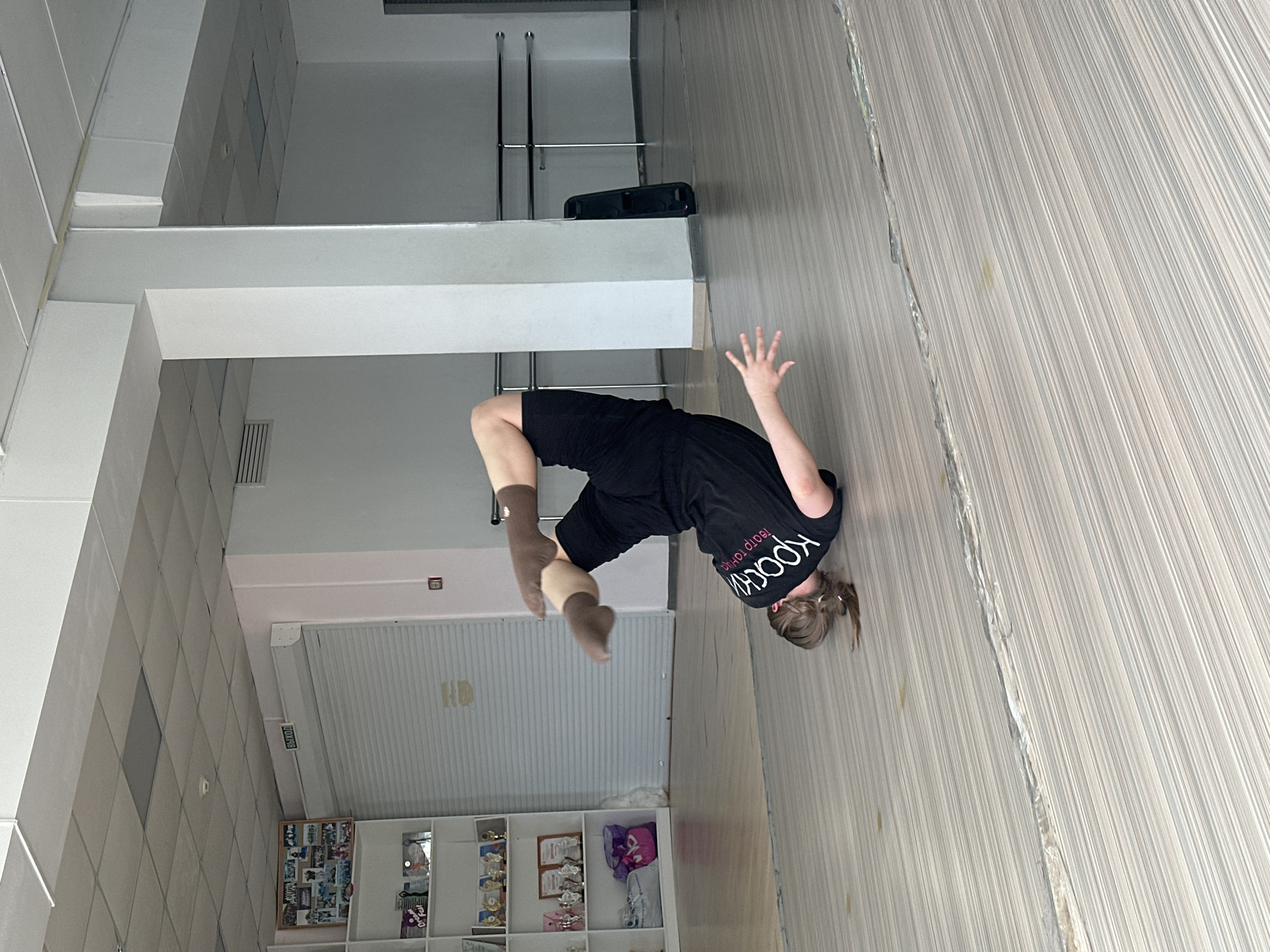 A woman in a black t-shirt and black shorts is in mid-air performing a flip inside a dance studio. The studio is located in Votkinsk, Russia, based on the Cyrillic text on her shirt. The walls and floors are predominantly white and gray. There is shelving along one wall filled with various items. There's a white support beam in the middle of the studio space. There is also exercise equipment behind her.