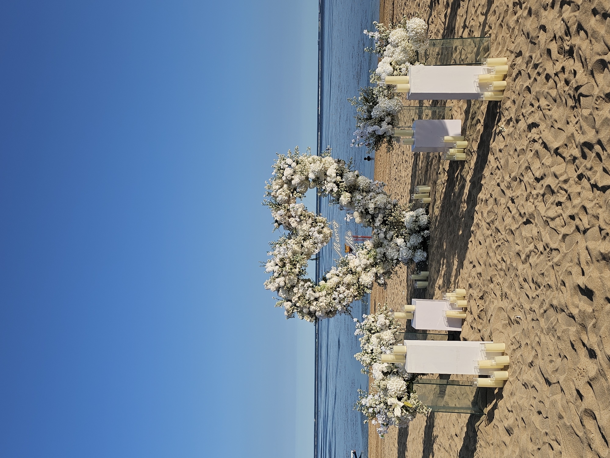 The image shows a wedding setup on a beach in Denpasar, Indonesia. A heart-shaped floral arch stands on the sand, facing the ocean. Flanking the arch are white platforms with cylindrical candles and floral arrangements. The sand is textured with footprints. The sky is clear and blue, with a visible horizon where the ocean meets the sky.