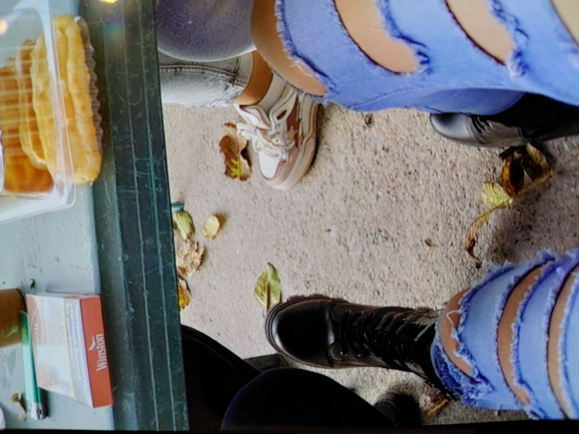 The image shows a ground-level view of a scene in Sofia, Bulgaria. There are three people present, identifiable only by their lower bodies. The person on the upper left wears light-wash jeans and sneakers. The person on the upper right wears distressed blue jeans and dark flats. The person on the lower right wears distressed blue jeans and black boots.
Located near the people is a partially eaten container of baked goods, a Winston cigarette pack, and a green lighter resting next to a dark green metal object. Autumn leaves are scattered on the ground.