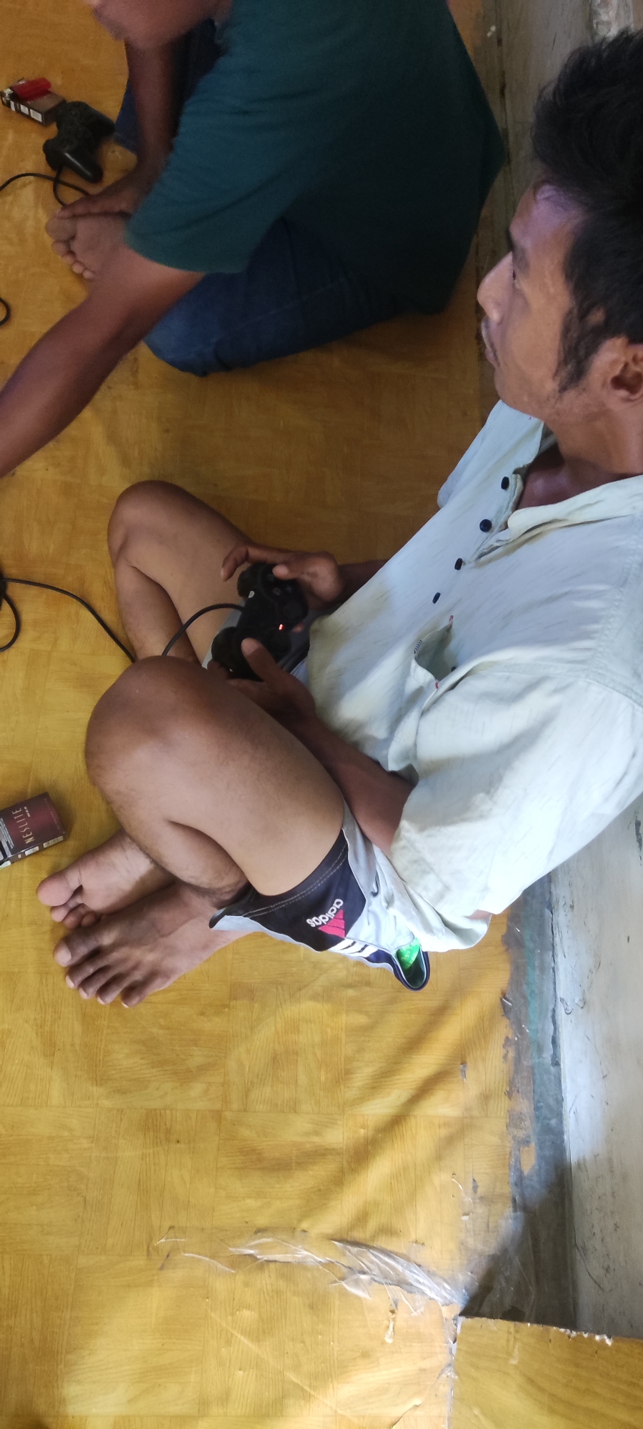 The image depicts two individuals, likely in a domestic setting in Karangwangun, Indonesia. In the foreground, a person is seated on a yellow linoleum floor, holding a black video game controller. They are wearing a light-colored, short-sleeved button-up shirt and athletic shorts with the Adidas logo visible. Their legs are crossed, and their bare feet are visible. To the left, a pack of cigarettes is placed on the floor. In the background, another individual is seated, wearing a dark green t-shirt and blue jeans, with their arm resting on their leg. The environment appears to be simple, with the floor being the dominant feature, exhibiting a wood-grain pattern. The lighting suggests an indoor space, possibly during daytime.