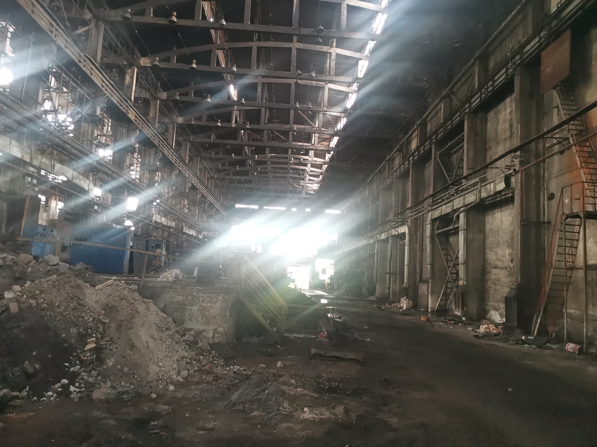 The image depicts the interior of a derelict industrial building, characterized by a high ceiling with exposed metal beams and light streaming through openings. The space is filled with debris and dirt, including a pile of rubble and scattered bricks on the floor. The walls are made of concrete and show signs of age and wear, with visible pipes and ladders attached to them. The scene suggests a state of abandonment and disuse. The specific location in Zhivkovo, Bulgaria, is not visually discernible.