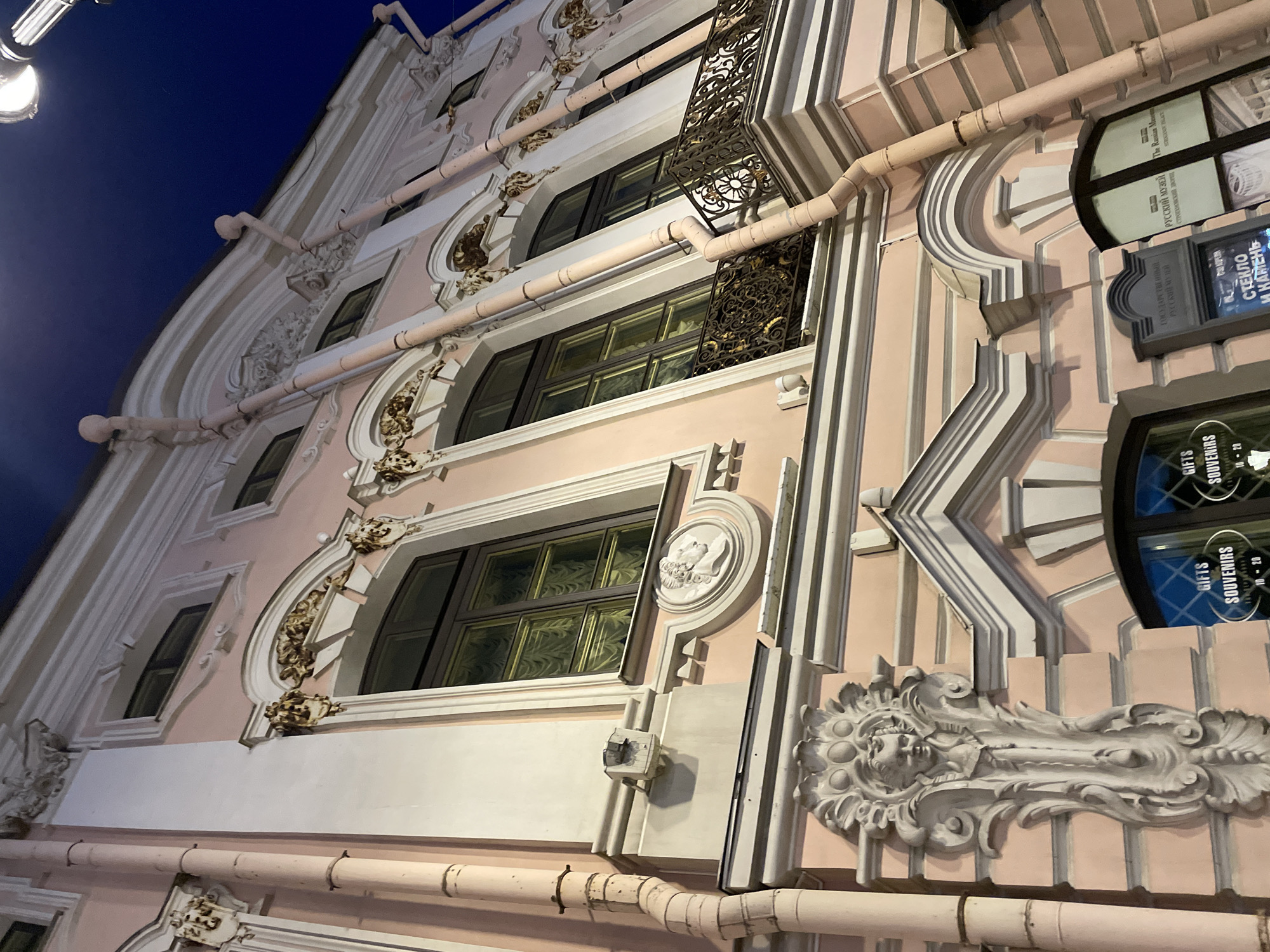The media file presents a dramatic, low-angle view of an ornate building facade at night or late dusk. The building exhibits a Baroque or Rococo architectural style, featuring soft peach-pink walls adorned with extensive white stucco or carved stone decorations. These intricate details include swirling motifs, floral patterns, and sculpted elements, such as a prominent bust on the lower right and smaller decorative forms around the windows.
Multiple windows, some with arched tops and others rectangular, are visible, featuring dark frames and, in some cases, ornate golden metallic grilles or balconies. Contrasting with the historical opulence are numerous light-colored pipes running across the facade. The deep, dark blue sky in the background, along with a partially visible bright street lamp in the upper left, confirms the nighttime setting.
No people are depicted, allowing the focus to remain on the architectural grandeur. Visible text on signs provides key information:
- "GIFTS SOUVENIRS 10 - 20" (repeated, indicating shop hours).
- "ГОСУДАРСТВЕННЫЙ РУССКИЙ МУЗЕЙ СТРОГАНОВСКИЙ ДВОРЕЦ" (The State Russian Museum Stroganov Palace) and its English translation "The Russian Museum STROGANOFF PALACE," clearly identifying the building as the Stroganov Palace, part of the Russian Museum.
- "СТЕКЛО И КАМЕНЬ" (Glass and Stone).
The image thus depicts a historic, elaborately decorated building, likely housing shops and a museum, under artificial lighting at night.