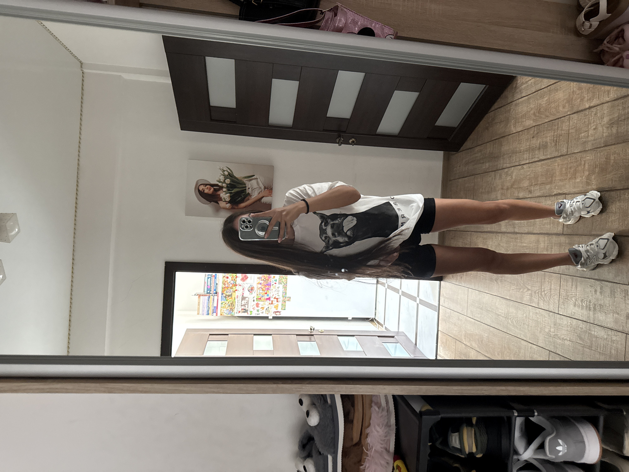 A young woman stands in front of a mirror, taking a selfie in what appears to be a room in Chișinău, Moldova. The room has light-colored walls and wooden flooring with a tiled pattern at the entryway. To the left, a tall mirror reflects the woman and part of the room. On the left side of the mirror, a shoe rack is visible, holding several pairs of footwear, including fuzzy slippers and athletic shoes.
The woman is wearing a white t-shirt with a graphic of a dog on it and black shorts. She has long, dark hair and is holding a smartphone in her right hand, angled to capture the photo. Her left hand is resting by her side.
Behind her, to the right, is a dark wooden door with rectangular glass panels. On the wall next to the door, there is a framed picture of a woman holding a bouquet of white tulips and wearing a wide-brimmed hat. The wall also has several colorful pictures or posters attached. A portion of a closet is visible on the far right. The overall impression is a casual, indoor setting, likely a bedroom or hallway.
