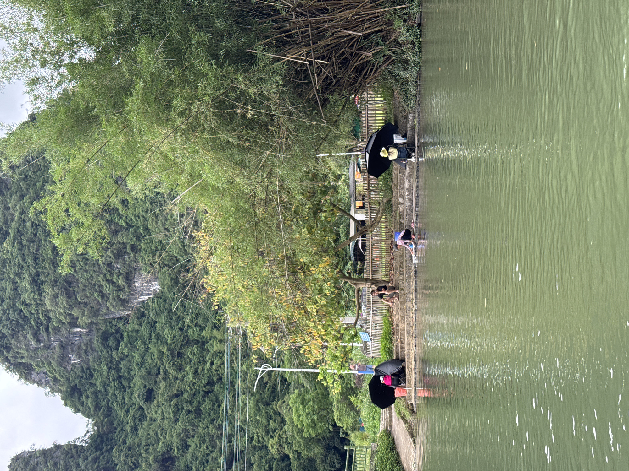 The scene depicts a body of water bordered by a bank or embankment in Guilin, China. Along the embankment, people are engaged in various activities: some are sitting or resting, possibly fishing or simply enjoying the view, while others appear to be wading or washing in the water. Some individuals are using umbrellas for shade. There are trees along the bank. In the background, a steep, forested hillside or mountain rises into the sky. There is an area with bamboo near the top right of the image.