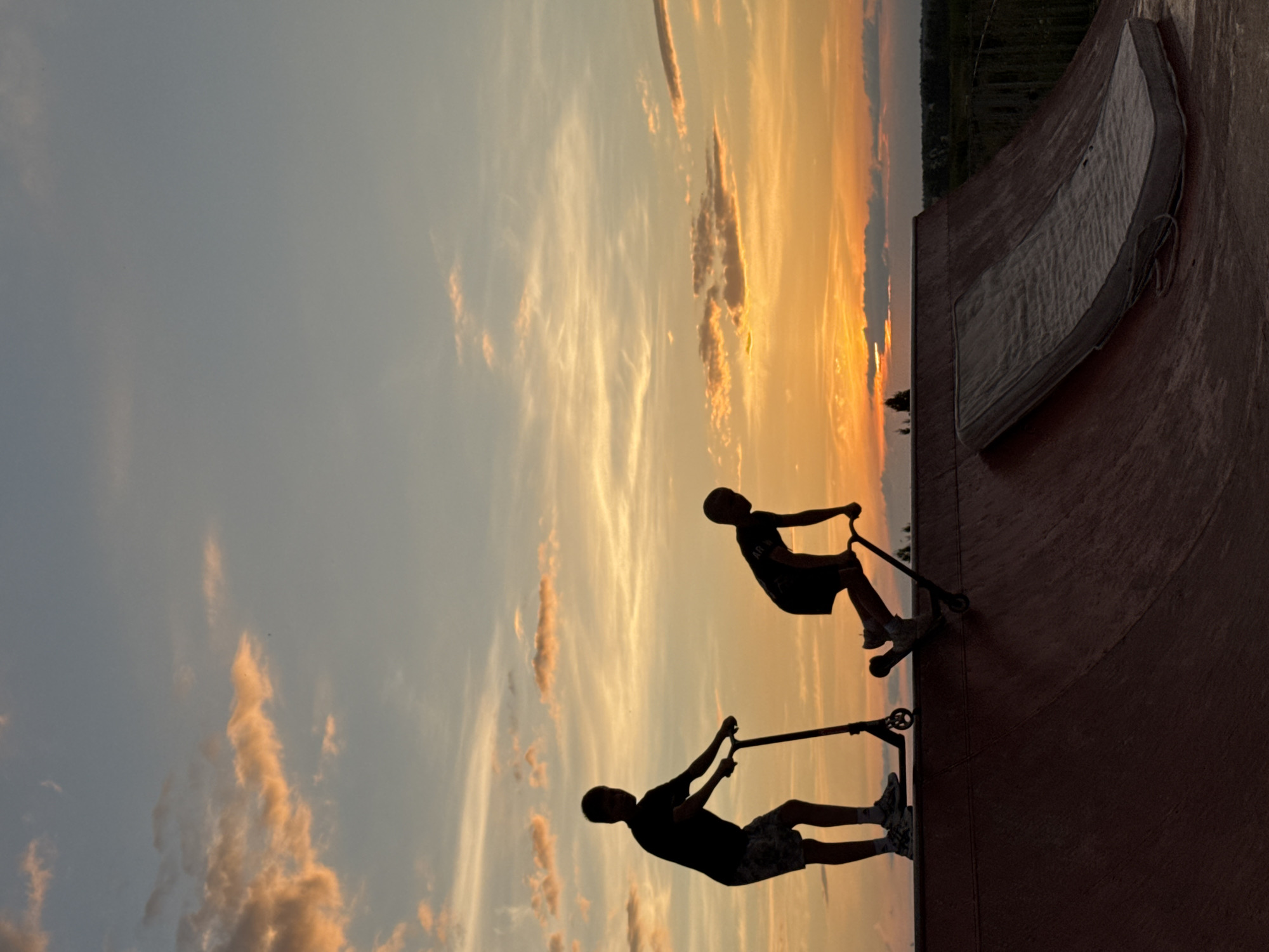 The image depicts a skate park at sunset in Mikhaylovsk, Russia. Two boys are silhouetted against the colorful sky, each on a scooter. One boy stands on the edge of a ramp, while the other is positioned further down, appearing to be in motion. A discarded mattress lies on the ramp surface. The sky displays a gradient of orange, pink, and blue hues, characteristic of a sunset. The foreground is dominated by the dark, curved surfaces of the skate park's ramps.