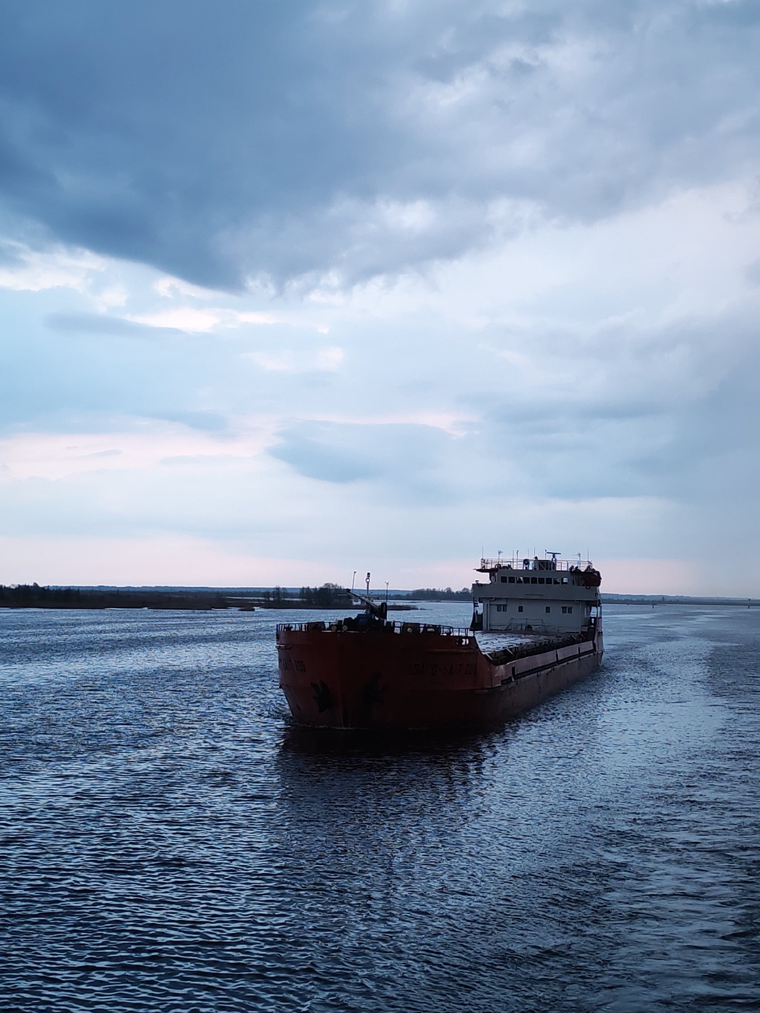 A cargo ship is sailing on the water. The photo was taken in Novokemskii, Russia. The sky is cloudy, with shades of blue and white. The water has a rippled surface and is reflecting the sky. The cargo ship is red and gray and is sailing away from the viewer. There is some land visible on the horizon.