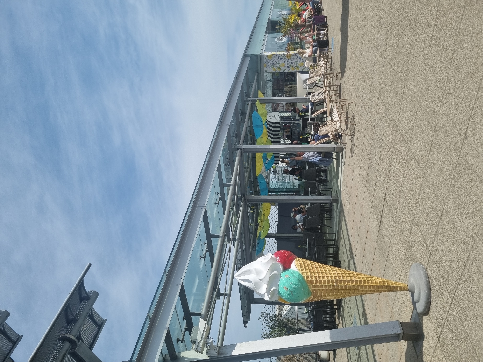 The image shows a life-sized decorative ice cream cone sculpture in front of a building in Echterdingen, Germany. The cone has a textured yellow waffle pattern and is topped with three scoops of ice cream: white, teal, and red. The sculpture is placed on a small gray stand. Behind the sculpture is a modern glass-walled building with tables and chairs inside, suggesting a restaurant or café. People are seated at some of the tables. Yellow and blue umbrellas are visible under the building's awning. The sky is blue with some clouds. The ground is paved with square tiles. Part of a structure with gray metal screens is visible on the left edge of the frame.
