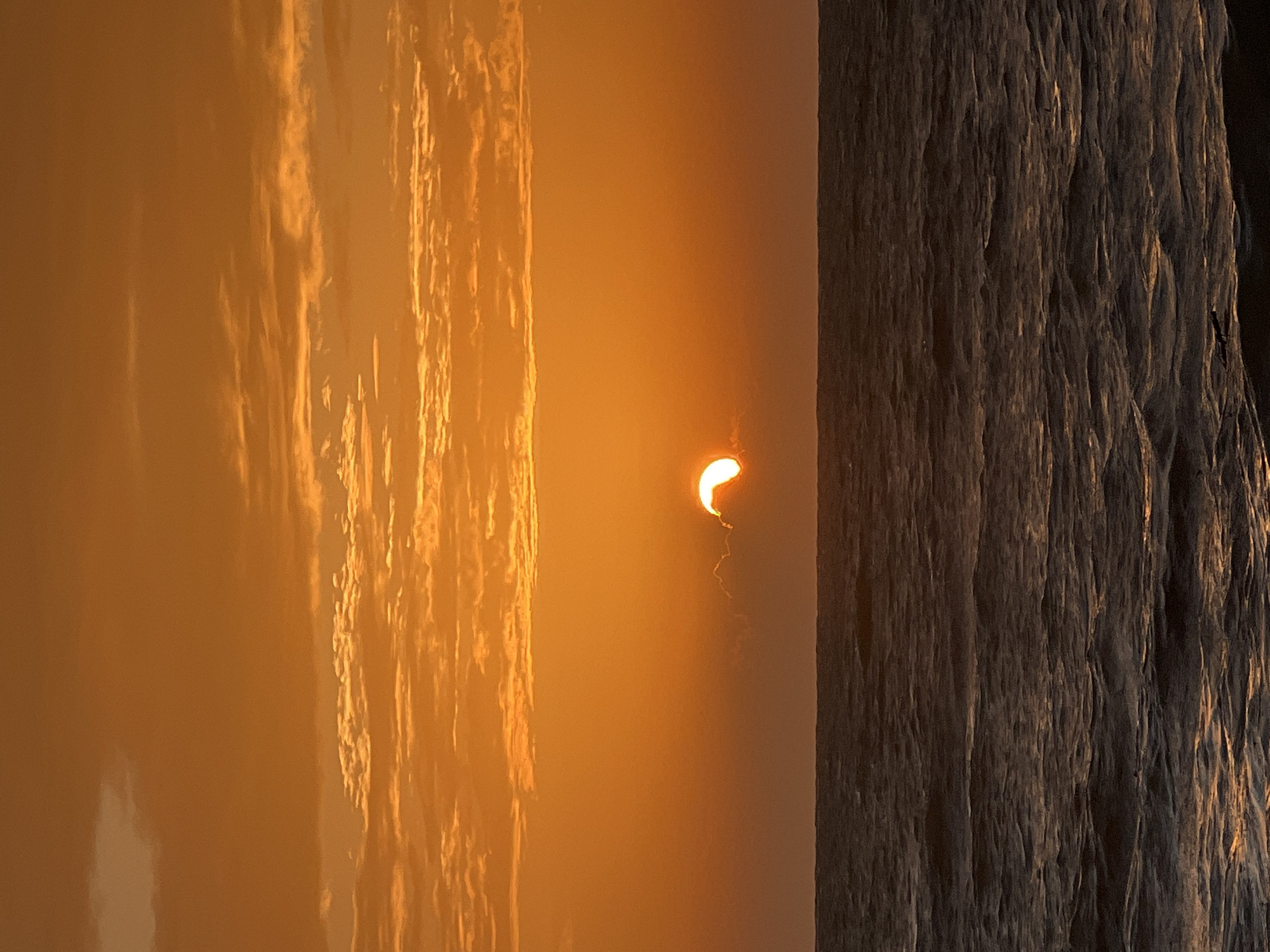 The image depicts a vibrant sunset over the ocean. The sky is a rich gradient of orange, with scattered clouds illuminated by the setting sun. A portion of the sun is visible, peeking through a thin cloud layer just above the horizon. The ocean surface is dark and choppy, with small waves reflecting the warm tones of the sky. The scene suggests a serene coastal environment. The location is described as being in Ko Pha-ngan District, Thailand. There are no people, animals, or man-made structures visible in the image.