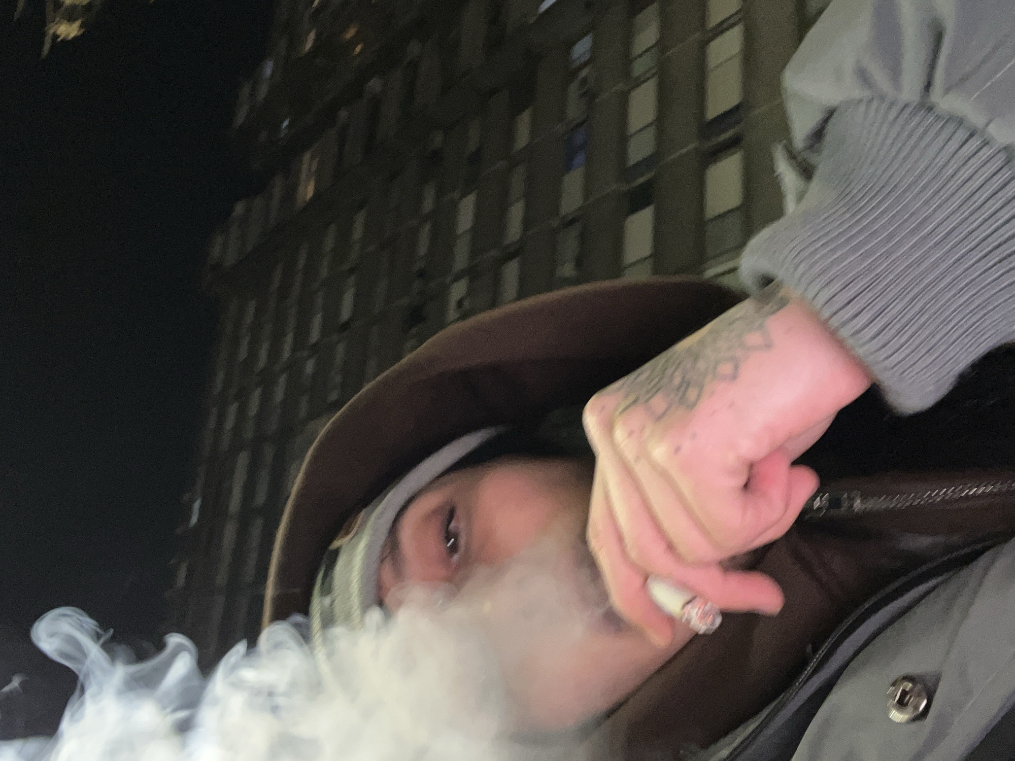 A low-angle shot shows a person smoking in an urban environment. The person, whose face is partially obscured by smoke, wears a brown hooded jacket and a grey shirt with ribbed sleeves. Tattoos are visible on the person's hand. They are holding a lit cigarette, exhaling a cloud of smoke. In the blurred background is a tall building illuminated by lights. The image was likely taken at night. It is not possible to determine the location is in Buenos Aires, Argentina from the provided image.