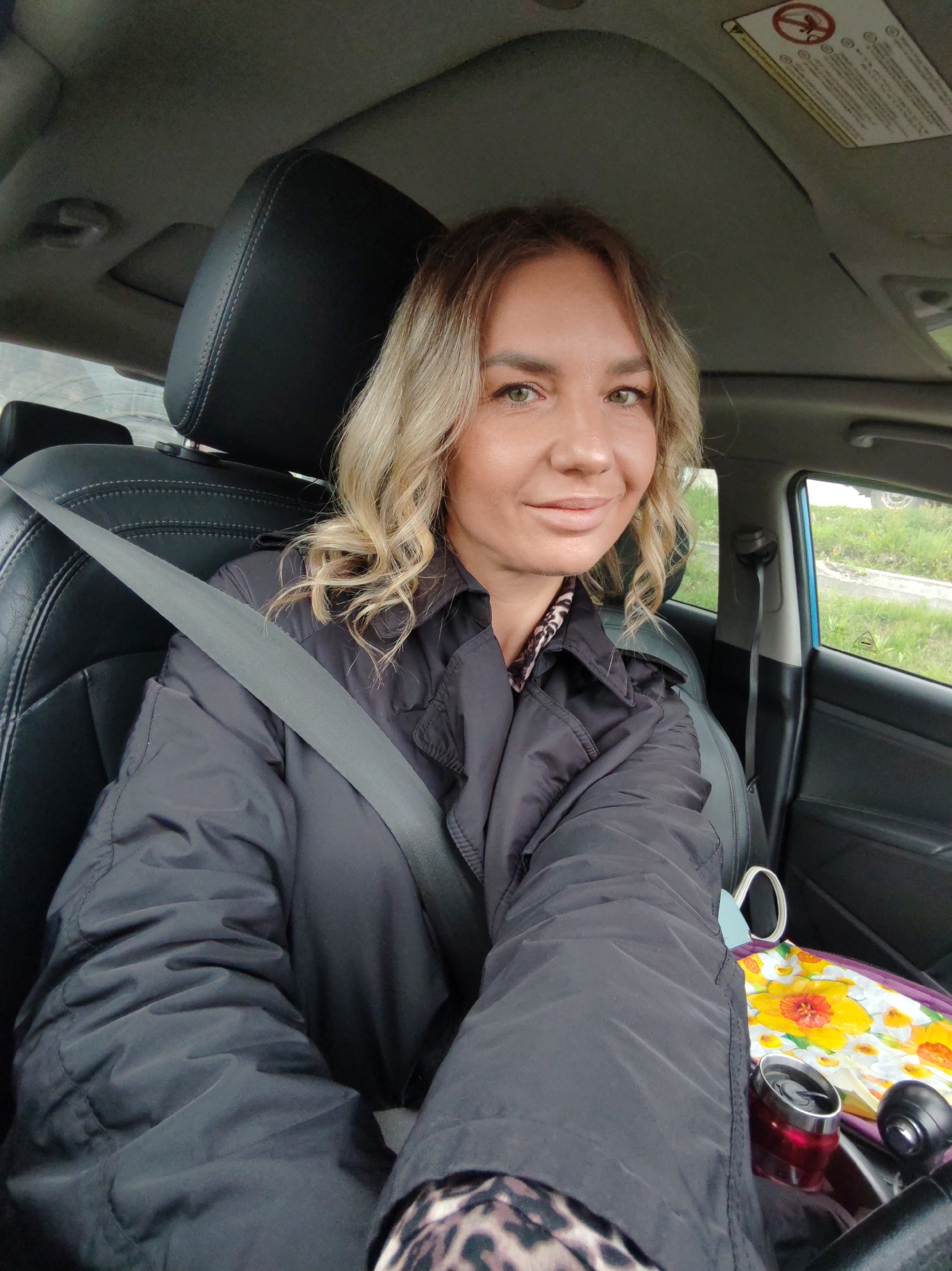 A woman is featured in a close-up selfie taken from inside a car, appearing to be in the passenger seat. She has blonde, wavy hair and light-colored eyes, looking directly at the camera with a gentle, pleasant smile. She wears a dark, possibly black or charcoal grey, puffer-style jacket, under which a leopard print pattern is visible at her neckline and wrist. A grey seatbelt is fastened across her chest.
The interior of the car is visible, featuring black leather seats with white stitching and a grey roof lining. Through the side window, green grass and a paved roadside are visible under what appears to be an overcast sky, suggesting it is daytime. On the sun visor, a safety warning label is clearly visible, displaying universal safety symbols and partially legible English text, including "WARNING" and references to "children" and "rear-facing child restraint," indicating airbag safety information.
Beside her, on the seat or console area, there are a few personal items: a vibrant bag or folded item with a yellow and orange floral pattern, and a red metallic thermos or travel mug. The scene conveys a casual and calm moment, likely while waiting or traveling in the car.