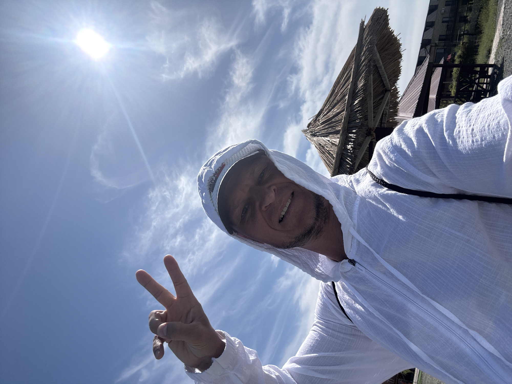 A man in a white hooded zip-up shirt and a white cap is taking a selfie outdoors under a bright, sunny sky with scattered clouds. He is smiling and holding up two fingers in a peace sign. In the background, there is a thatched umbrella, some gazebos, and a building. The image appears to be taken in Zharbulak, Kazakhstan.
