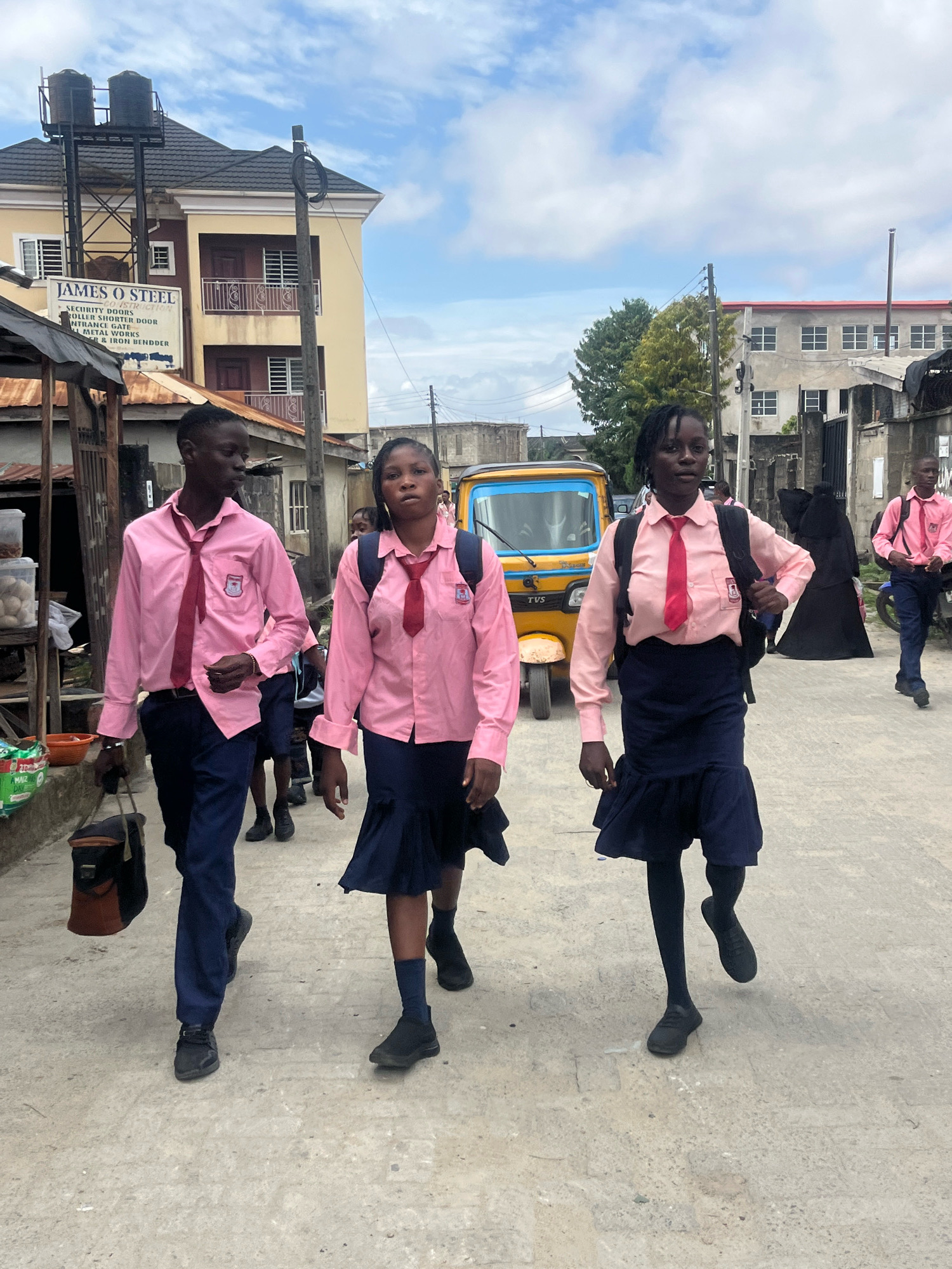 In Ado, Nigeria, a group of students walks along a concrete path. The students, three prominently in the foreground and others further back, are dressed in matching pink long-sleeved shirts and dark blue skirts or trousers, accented with red ties. They appear to be walking away from the camera. A young man in the front left carries a dark brown and black satchel. To their right, a yellow and blue three-wheeled vehicle, commonly known as a "keke," is visible. Behind the students, buildings line the street, including a two-story structure with a sign reading "JAMES O STEEL" and another building with a balcony. The sky above is partly cloudy. The overall scene depicts a typical day on a street in Ado, with students on their way somewhere.