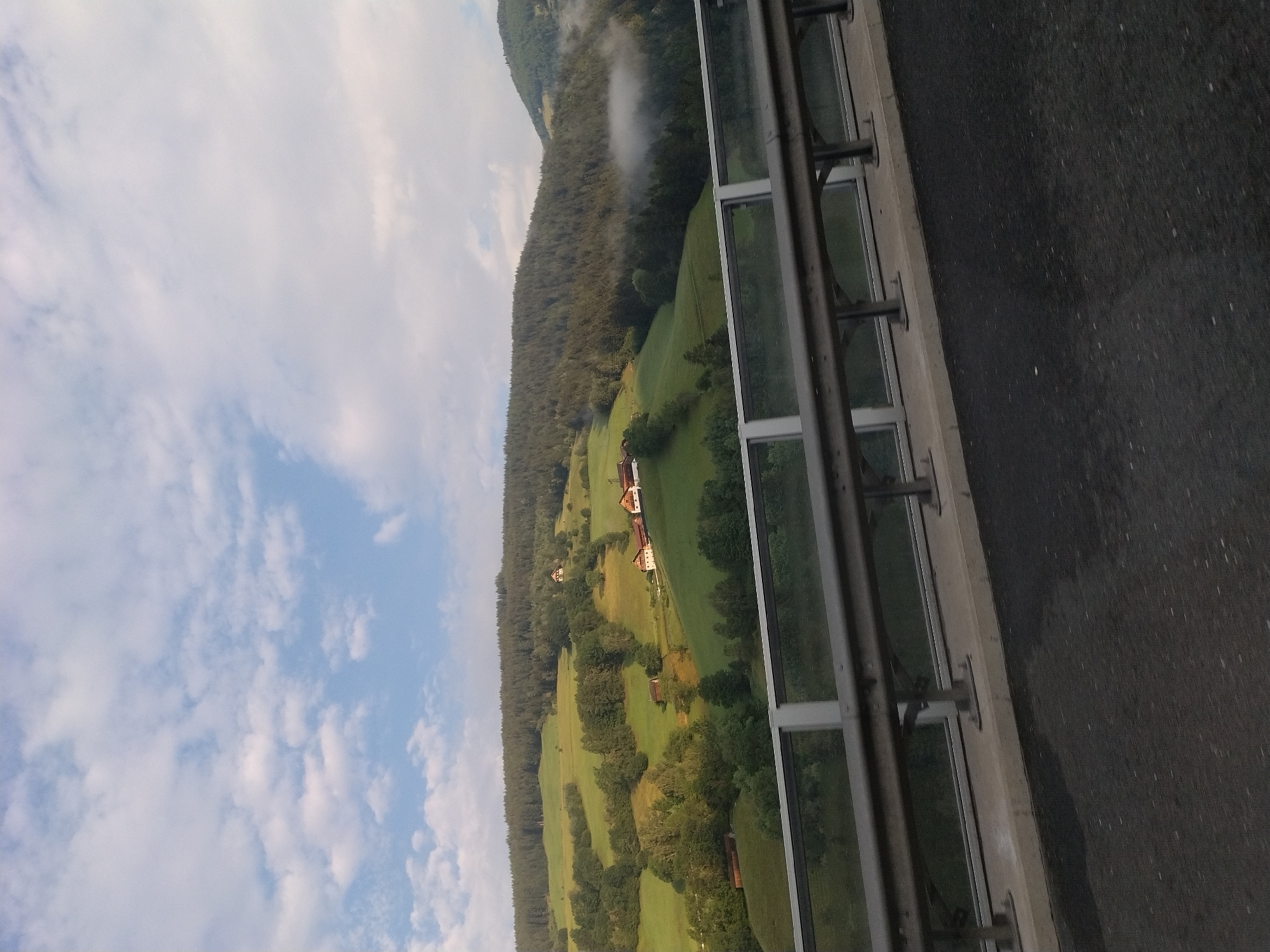 The image depicts a scenic view of rolling green hills and forested mountains under a partly cloudy sky. In the foreground, a modern guardrail with glass panels separates the viewer from the landscape. The road surface in the immediate foreground is dark asphalt. Scattered across the hills are several houses, some with red roofs, nestled amongst the greenery. A light mist or fog hovers in a valley in the background, partially obscuring the trees. The overall impression is of a rural or semi-rural setting, likely in a mountainous region. The location is identified as Krems in Kärnten, Austria. There are no people or animals visible in the image.