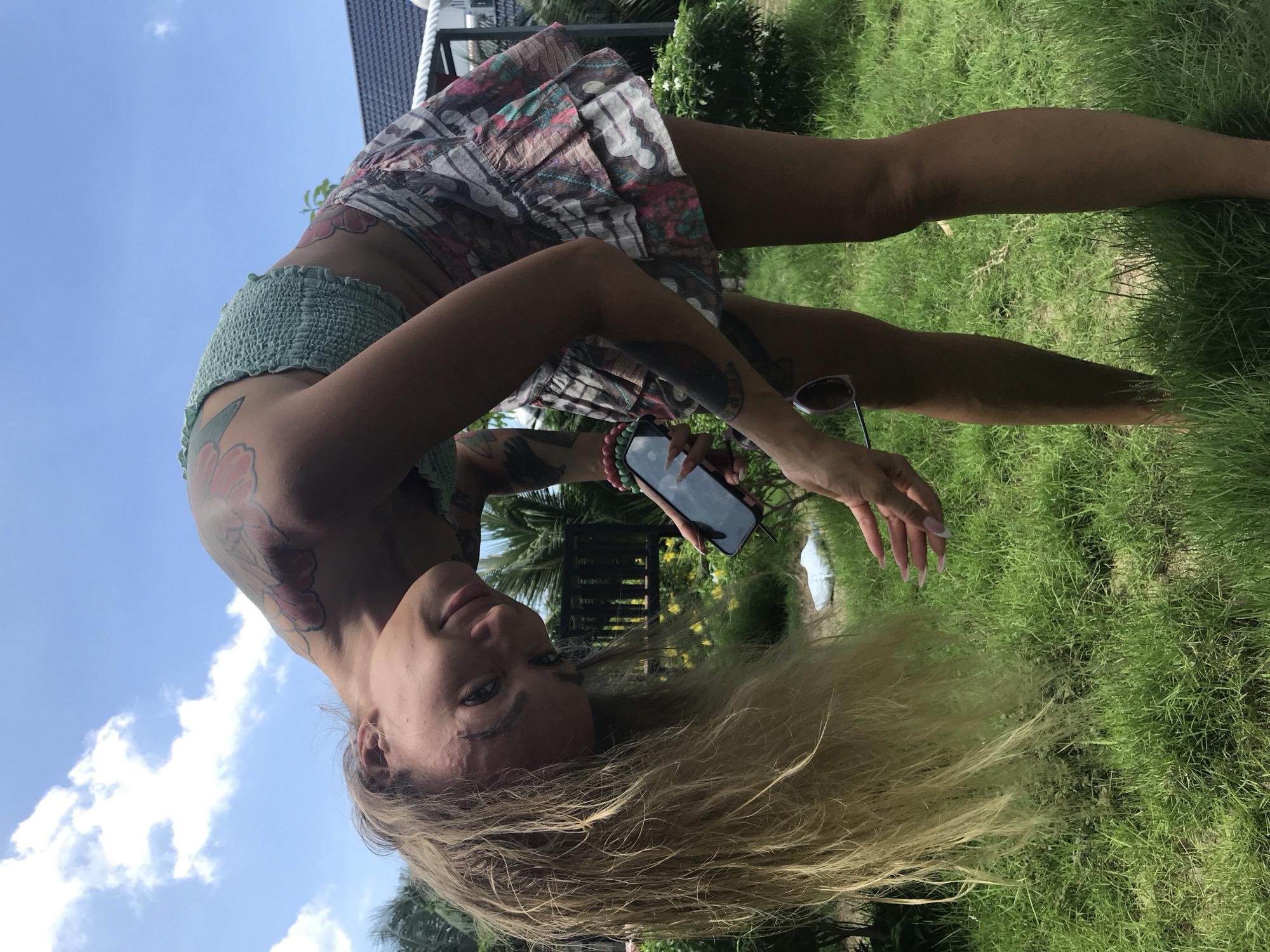 A female subject is captured in an outdoor, brightly lit setting in Mũi Né, Vietnam, under a clear blue sky with scattered white clouds. The subject is bending forward at the waist, with her head tilted towards the camera, presenting a faint smile. Her long, light-colored hair hangs towards the ground. She has a tanned complexion.
She is wearing a light green, textured, strapless crop top and a short, tiered skirt with a colorful floral and geometric pattern primarily in shades of pink, teal, brown, and white. Visible tattoos include a large floral design on her right shoulder/upper arm, another tattoo of a darker, possibly solid color on her left upper arm, and a partial floral design on her right side/lower back.
On her right wrist, she wears multiple beaded bracelets in red and green. Her right hand holds a black smartphone with a reflective screen; her thumb has a pink-tipped nail. Her left hand, with long nails featuring a French manicure (pink base, white tips), is positioned near her side, and a pair of sunglasses hangs from this hand.
The ground is covered in green grass, with some areas appearing sparse. A small white object resembling a plastic bag is visible on the grass to her left. The background features dense tropical foliage, including palm trees and other green plants, along with yellow flowering bushes. A building with a dark-tiled roof is visible in the upper right, accompanied by a white railing and a dark wooden fence structure further in the background.