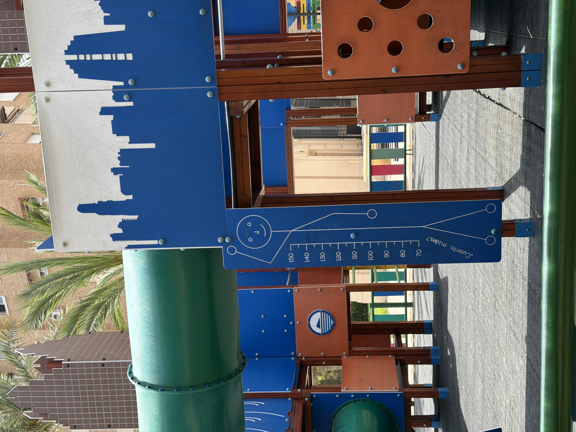 The image depicts a section of a colorful outdoor playground, likely designed for children. The structure is made of various materials, including blue and orange panels, wooden beams, and a large green cylindrical tube that appears to be a slide or tunnel. The playground elements are arranged in a multi-level design, with geometric shapes and cutouts visible.
One prominent blue panel features a stylized silhouette of a city skyline in white. Another blue panel displays a ruler-like graphic with numerical markings from 70 to 150, and a simple drawing of a stick figure with a smiley face. Text in Spanish, "¿Cuanto mides?", which translates to "How tall are you?", is visible along the bottom edge of this panel, indicating the graphic is a height measurement tool.
Other orange panels have circular cutouts, possibly for climbing or visual interest. A small circular emblem with a stylized boat or sail is present on one of these orange panels. Interspersed within the structure are wooden slats and supports, suggesting a sturdy construction.
In the background, hints of the setting are visible. A palm tree with its fronds is partially visible on the left side, suggesting a warm climate. Behind the playground structure, a building with windows and a tiled facade can be seen, along with other playground elements in muted colors. The ground appears to be a paved surface, possibly asphalt or concrete, with some visible cracks and shadows cast by the playground structure.
The overall impression is a bright, sunny day, given the clear shadows. The setting is an outdoor recreational space, consistent with a park or a schoolyard. The playground appears well-maintained and designed for active play. The image is taken from a slightly tilted perspective, adding a dynamic feel to the composition. There is no indication of people or any specific event taking place at the moment the photograph was captured. The location is identified as Benidorm, Spain.
