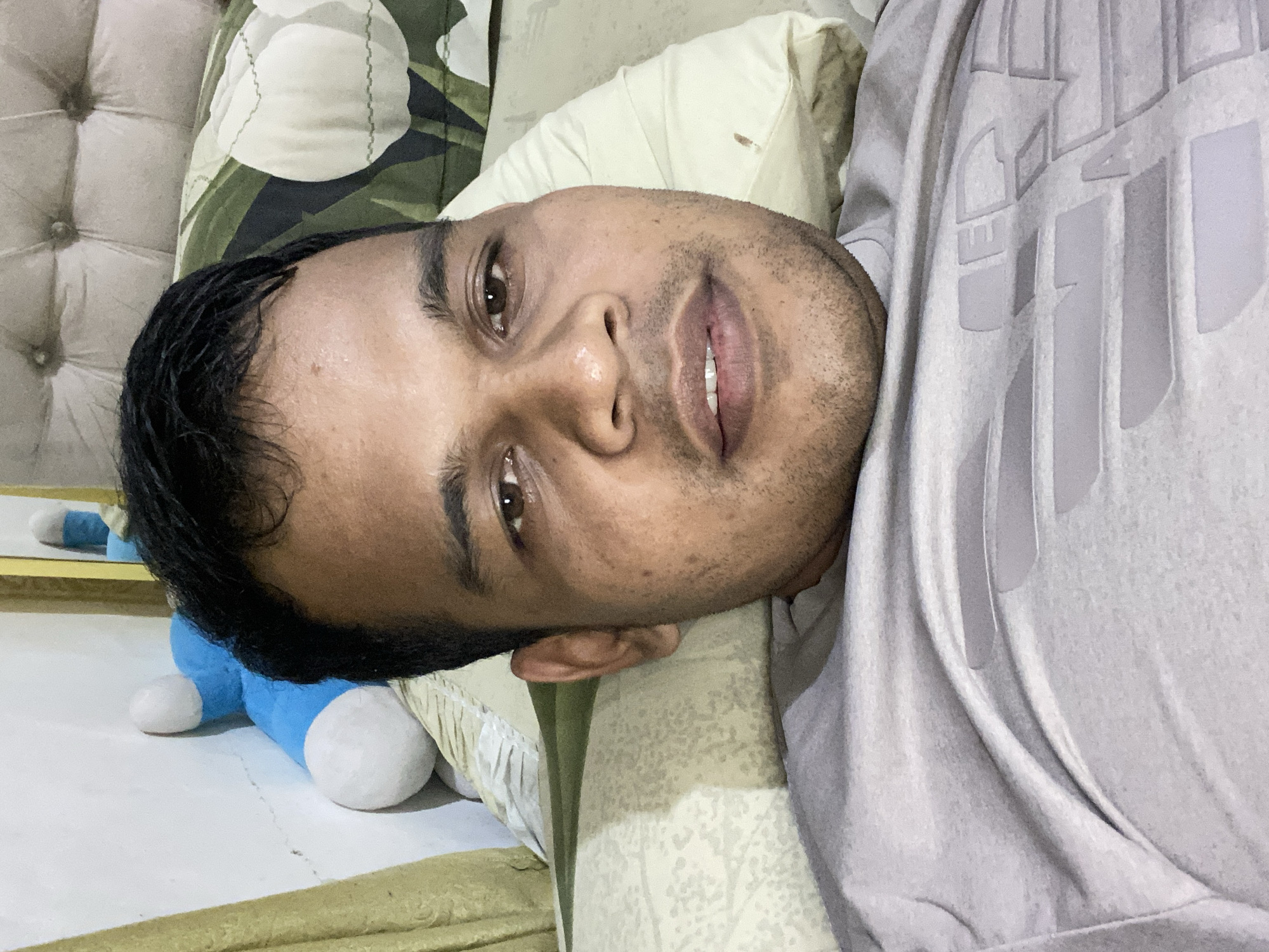 A close-up, selfie-style photograph shows a man lying down, propped up against pillows on a bed. He is wearing a light gray t-shirt with a large, embossed graphic on the front. The man has dark hair and brown eyes. In the background, to the left, a blue stuffed toy is visible leaning against a white wall, partially obscured by a yellow curtain. The bed has a textured headboard and patterned pillows. The lighting appears to be natural, coming from an unseen source. The overall setting suggests a bedroom environment. The image is taken from the perspective of someone lying on the bed, looking directly at the camera.