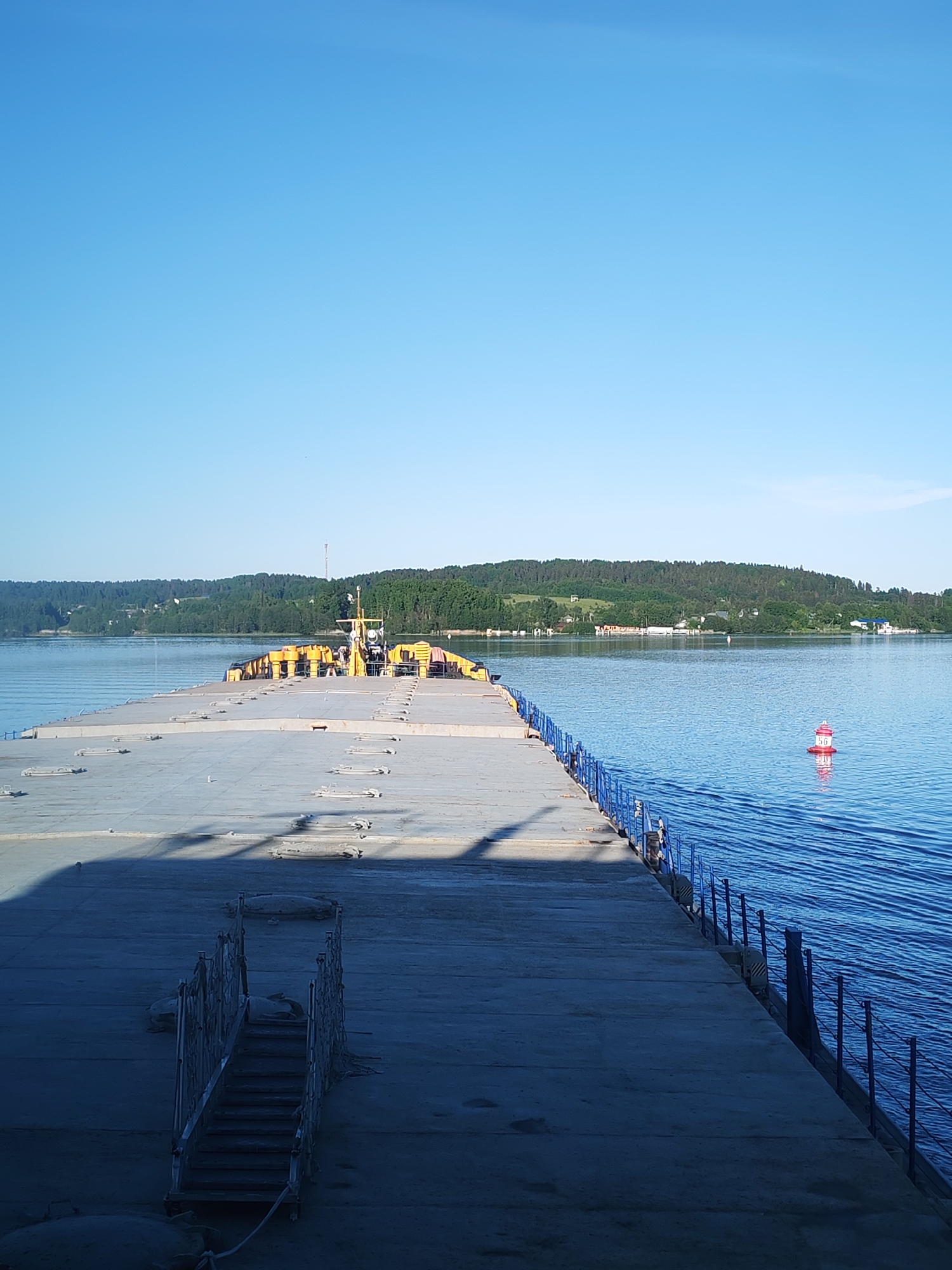 This image depicts a concrete pier extending into a calm body of water under a clear blue sky, located in Goritsy, Russia. The pier occupies the lower half of the frame, angled slightly to the right, creating a leading line towards the horizon. Its surface is light gray concrete, showing some wear and a distinct shadow extending from the left, indicating sunlight from the upper left. Numerous rectangular metal cleats or mooring points are embedded along its length. In the lower left foreground, a narrow set of dark metal stairs with ornate railings descends from the pier's edge.
Along the right side of the pier, a blue metal railing with vertical posts and horizontal bars runs its entire visible length. The water on the right is deep blue, exhibiting subtle ripples. Approximately mid-frame on the right, a red and white striped navigational buoy, cylindrical in shape with a rounded top, is visible, marked with the number "56".
Towards the far end of the pier, a section of yellow industrial equipment or a barge structure is docked or integrated. This yellow section features various mechanisms and what appears to be a control cabin or engine room. Beyond this, the water extends to a distant shoreline. The background consists of gently rolling hills densely covered with green trees and scattered patches of lighter foliage. A few small, distant buildings, including one prominent white structure, are visible among the trees on the far shore. The sky above is a uniform, bright light blue, with no clouds. No people are present in the scene.