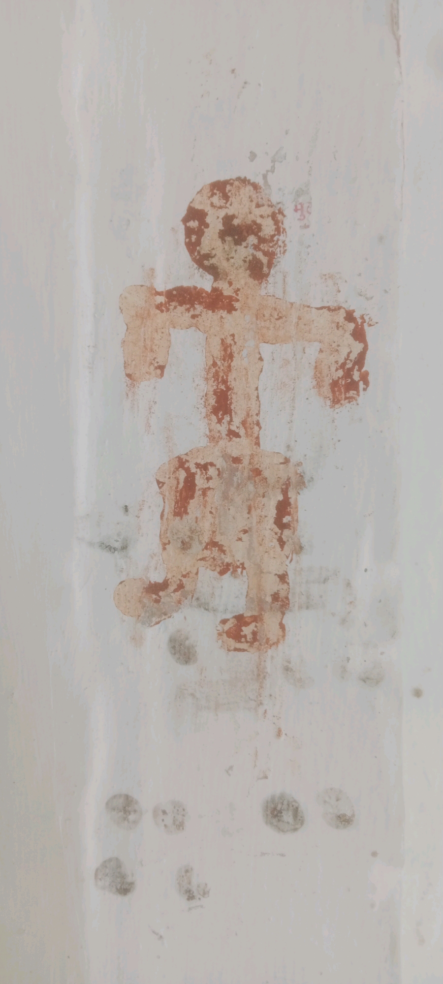 The image depicts a faded, crudely drawn red figure resembling a stick person on a white wall. The figure has a circular head, a rectangular torso, outstretched arms, and legs. The drawing appears to be ancient and weathered, with parts of the pigment chipped away, revealing the white background. Below the figure, there are several dark, circular smudges on the wall, possibly footprints or other markings. The background wall is not uniform, showing signs of wear and tear, with streaks and uneven texture. In the upper right corner, a faint pink inscription is visible, though illegible. The image is taken from a vertical perspective, with the figure centered and taking up most of the frame. The context indicates this is located in Ml#alankheri, India.